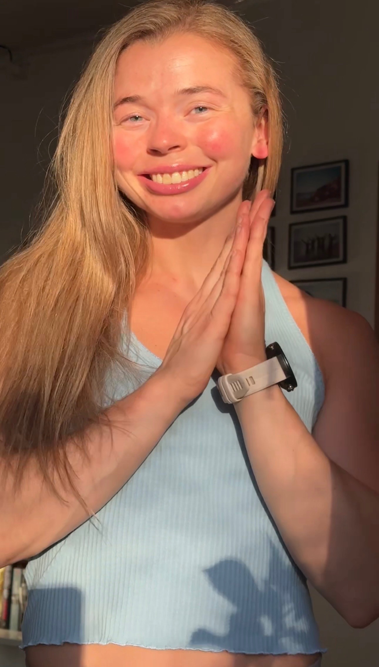 A smiling person with long hair, wearing a light blue tank top, poses with hands together in a warm, sunlit room.