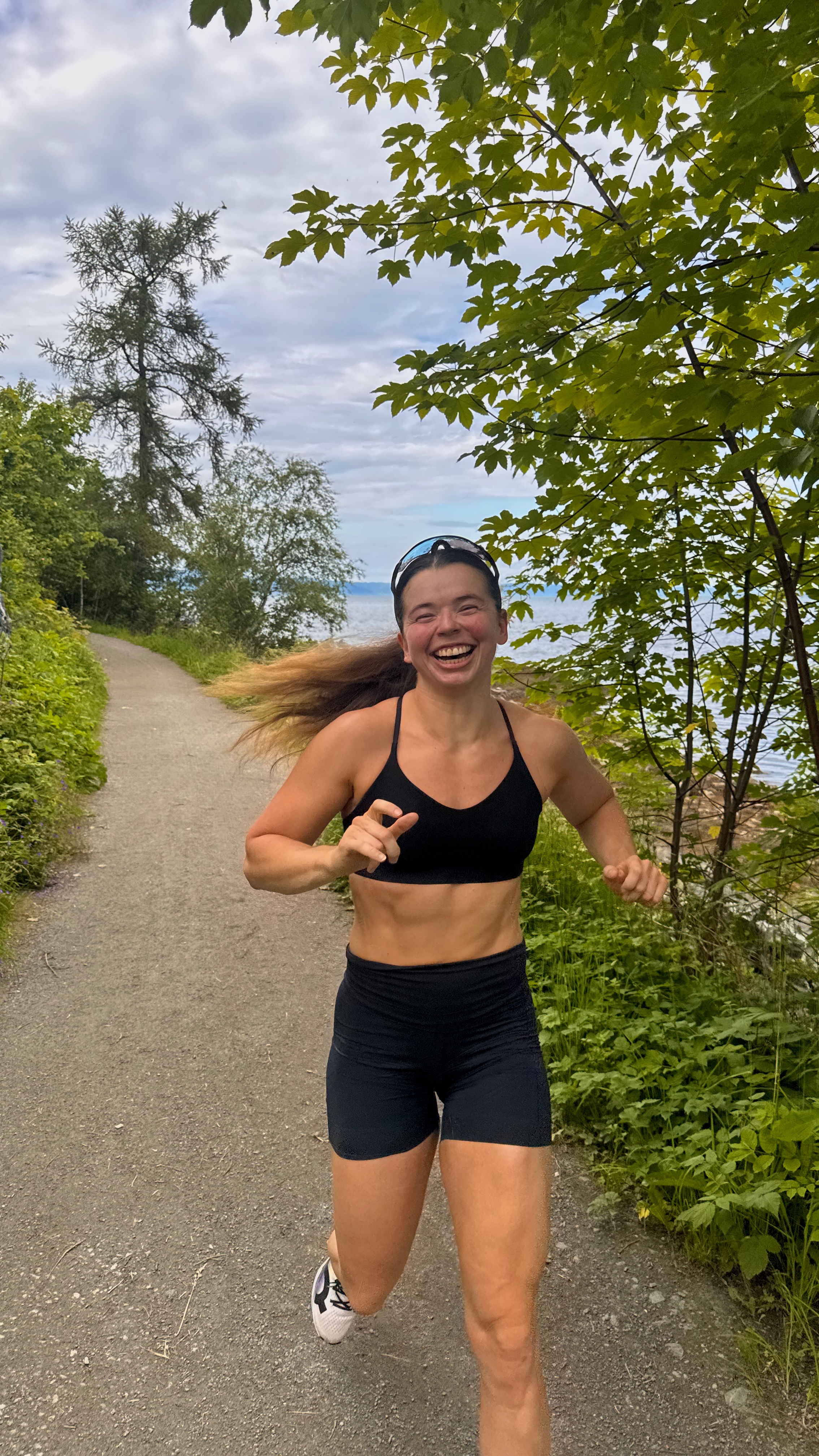 A woman in athletic wear runs along a scenic path surrounded by greenery and water, smiling joyfully.