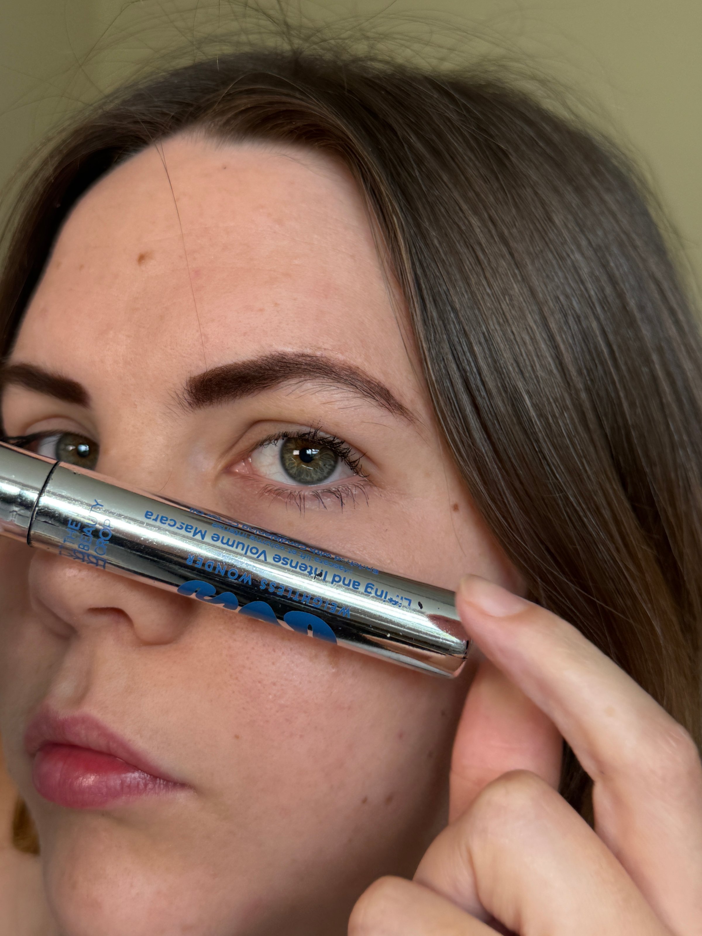 A close-up portrait of a woman holding a mascara tube near her eye, showcasing her makeup look.