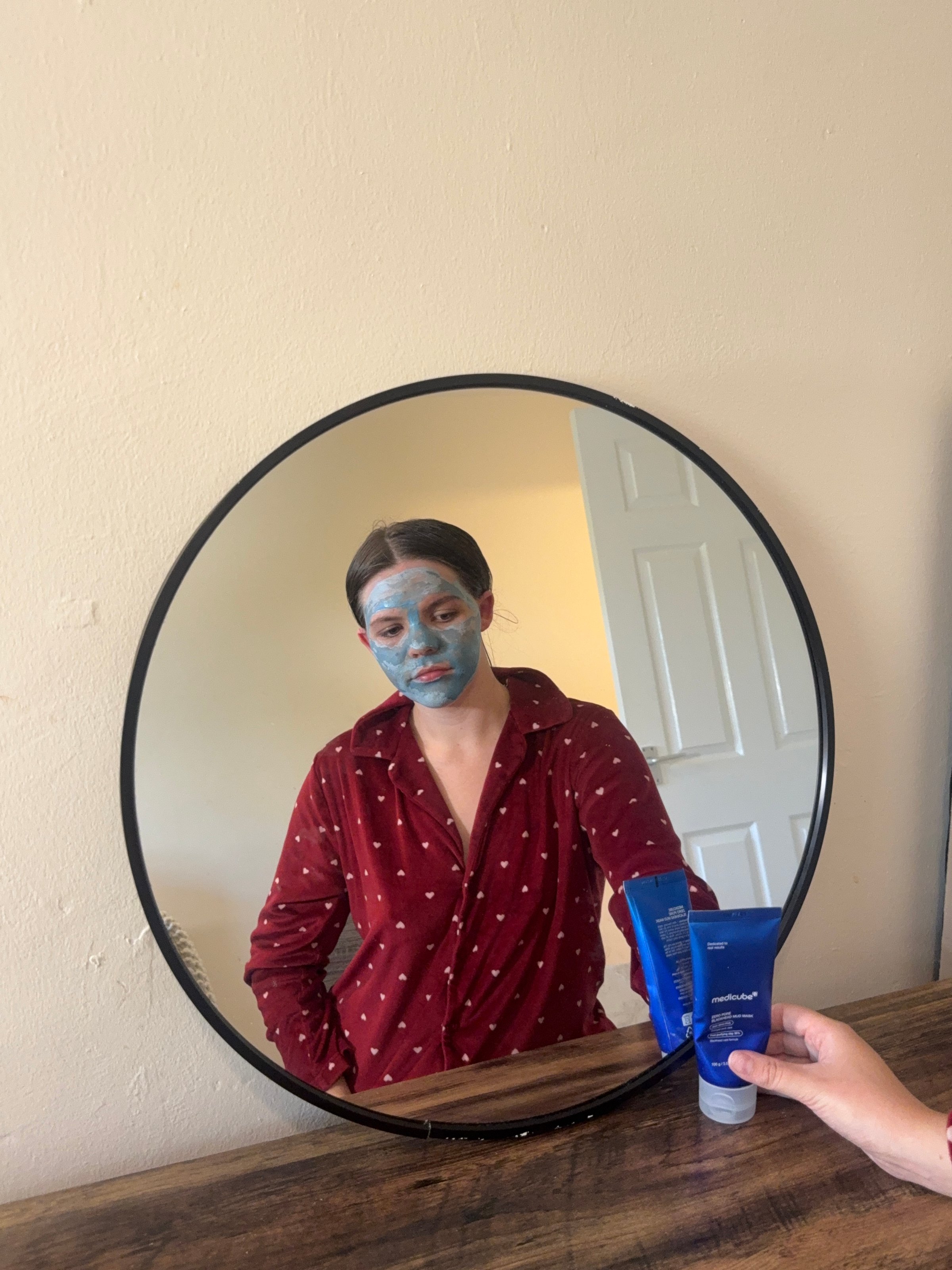 A person with a blue facial mask is reflected in a round mirror, wearing a red polka dot shirt and holding a skincare product.