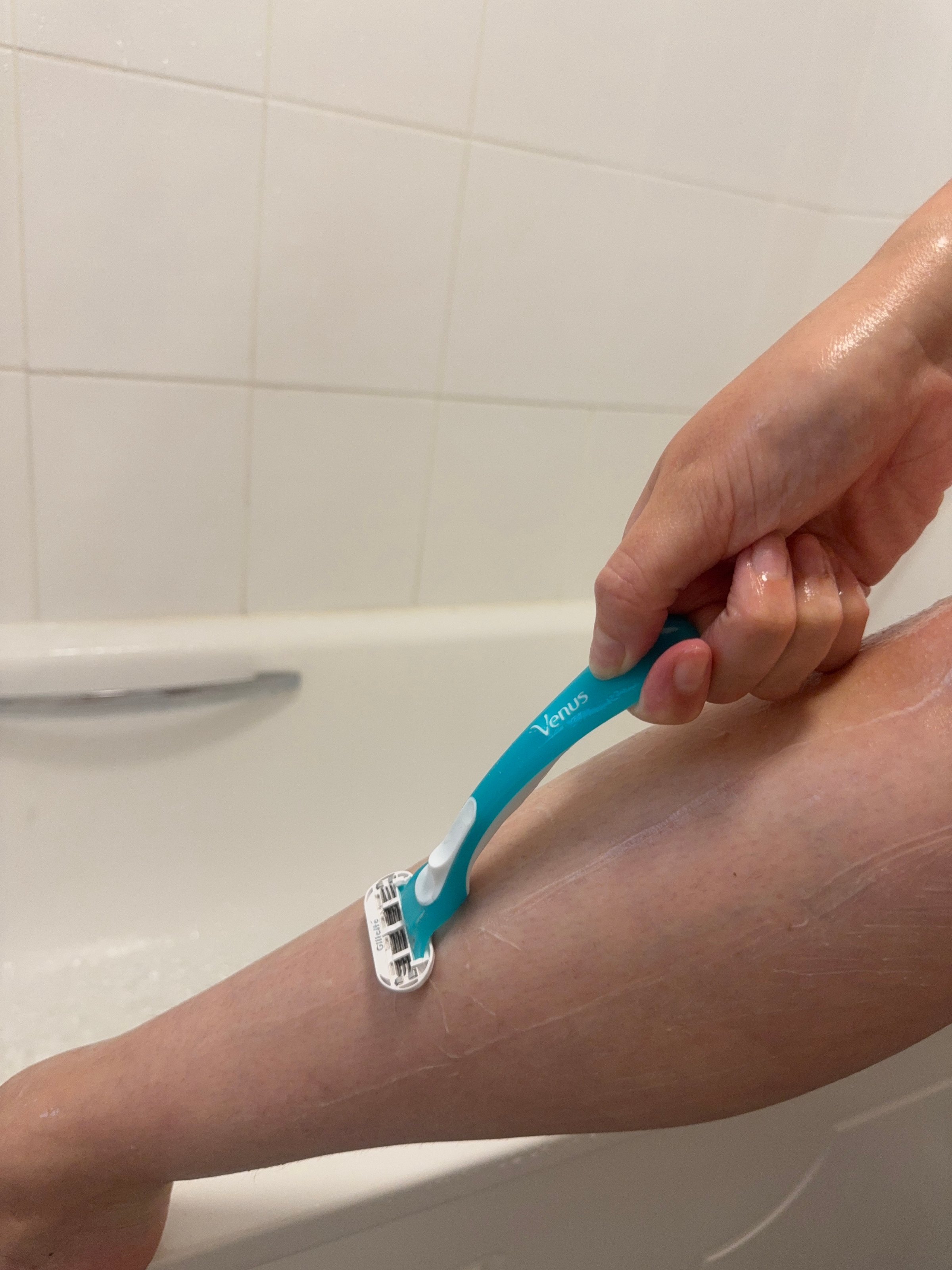 A close-up of a hand using a razor to shave a leg in a bathtub setting.