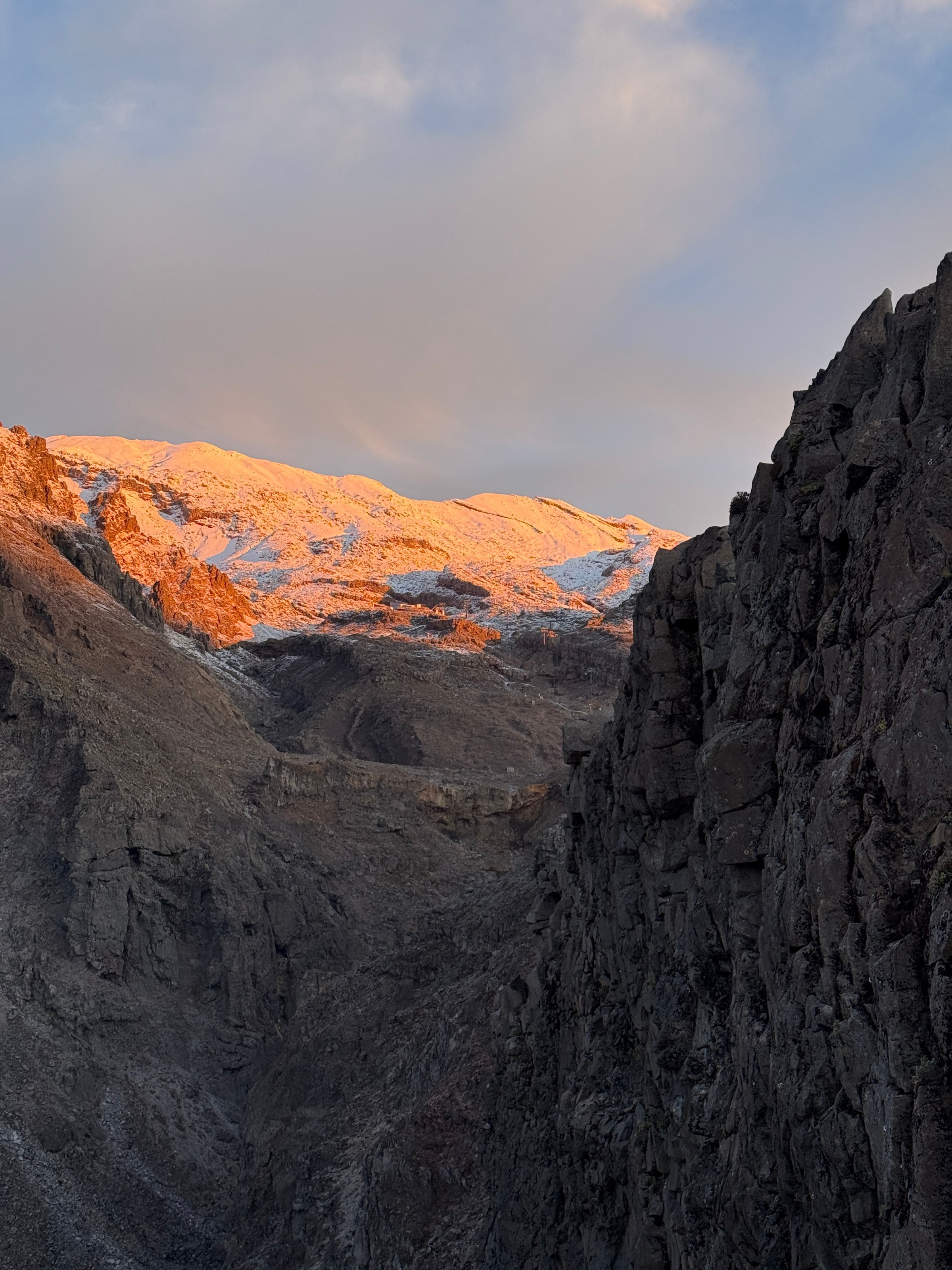 A breathtaking view of a mountain range illuminated by the warm glow of the setting sun, with rugged cliffs framing the scene.