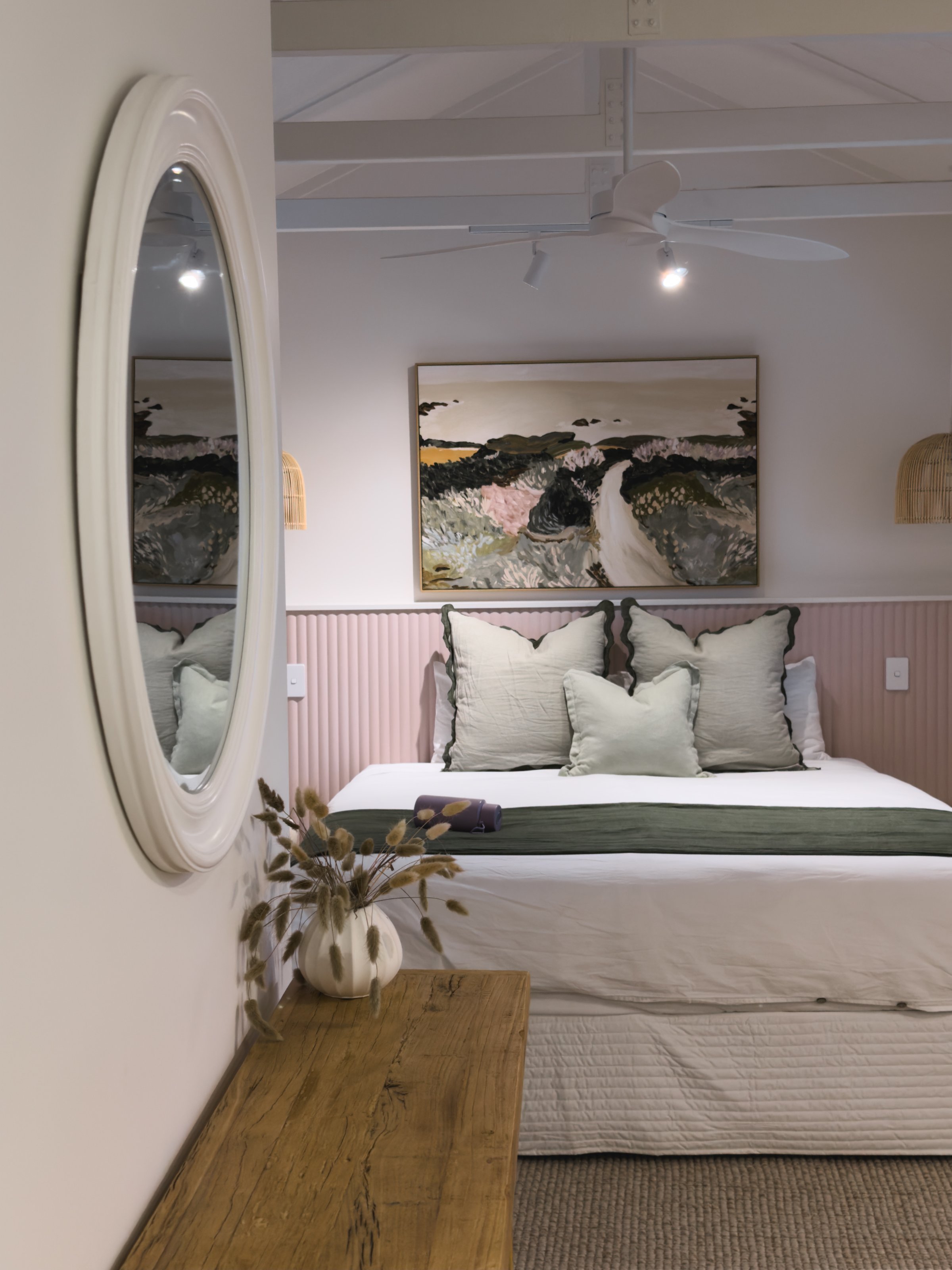 A cozy bedroom featuring a white bed with decorative pillows, a wooden dresser, and a round mirror reflecting a painting on the wall.
