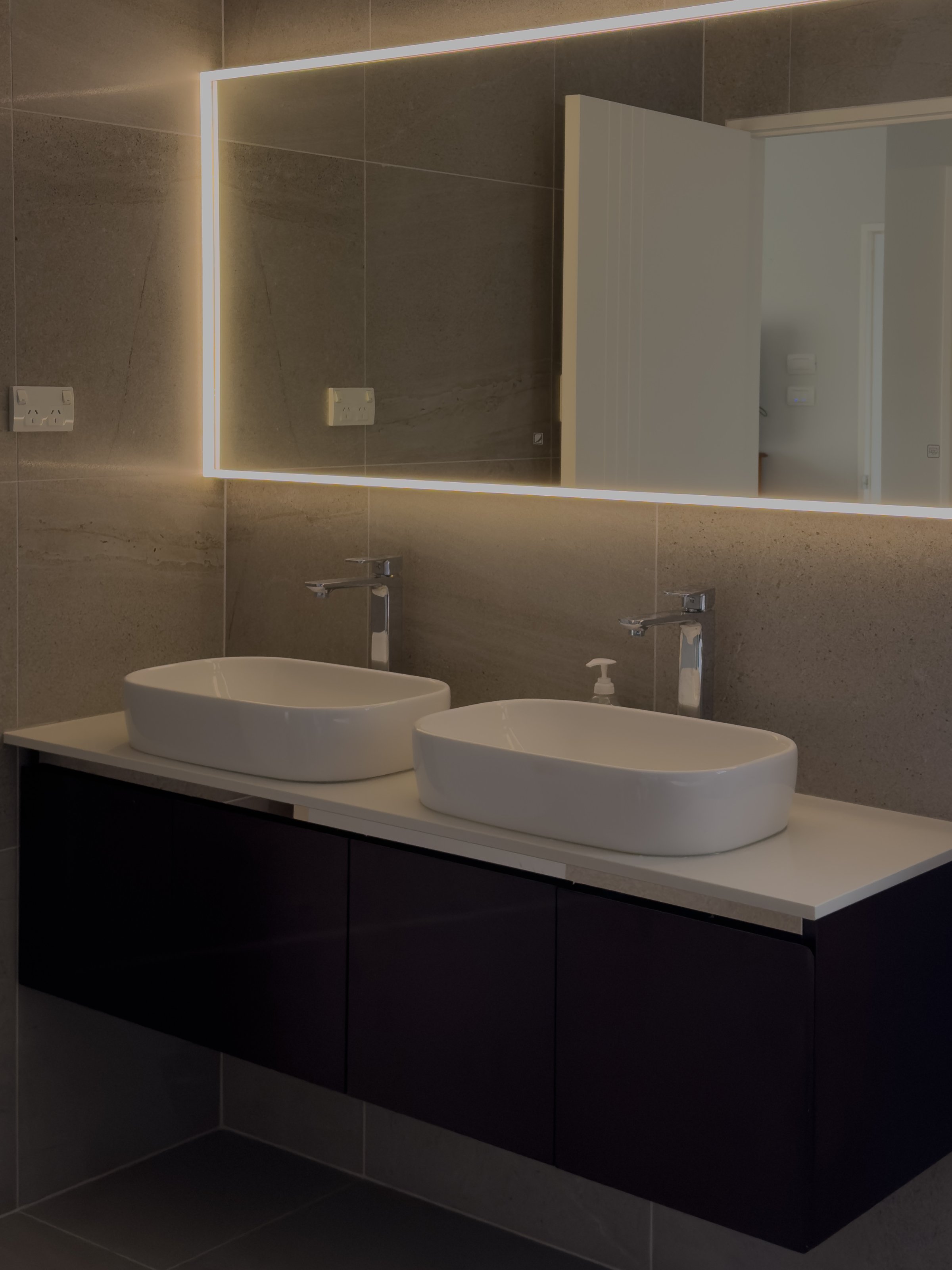 A modern bathroom featuring a sleek double sink with a minimalist design and a backlit mirror above. The walls are tiled in a neutral tone, creating a clean and contemporary aesthetic.