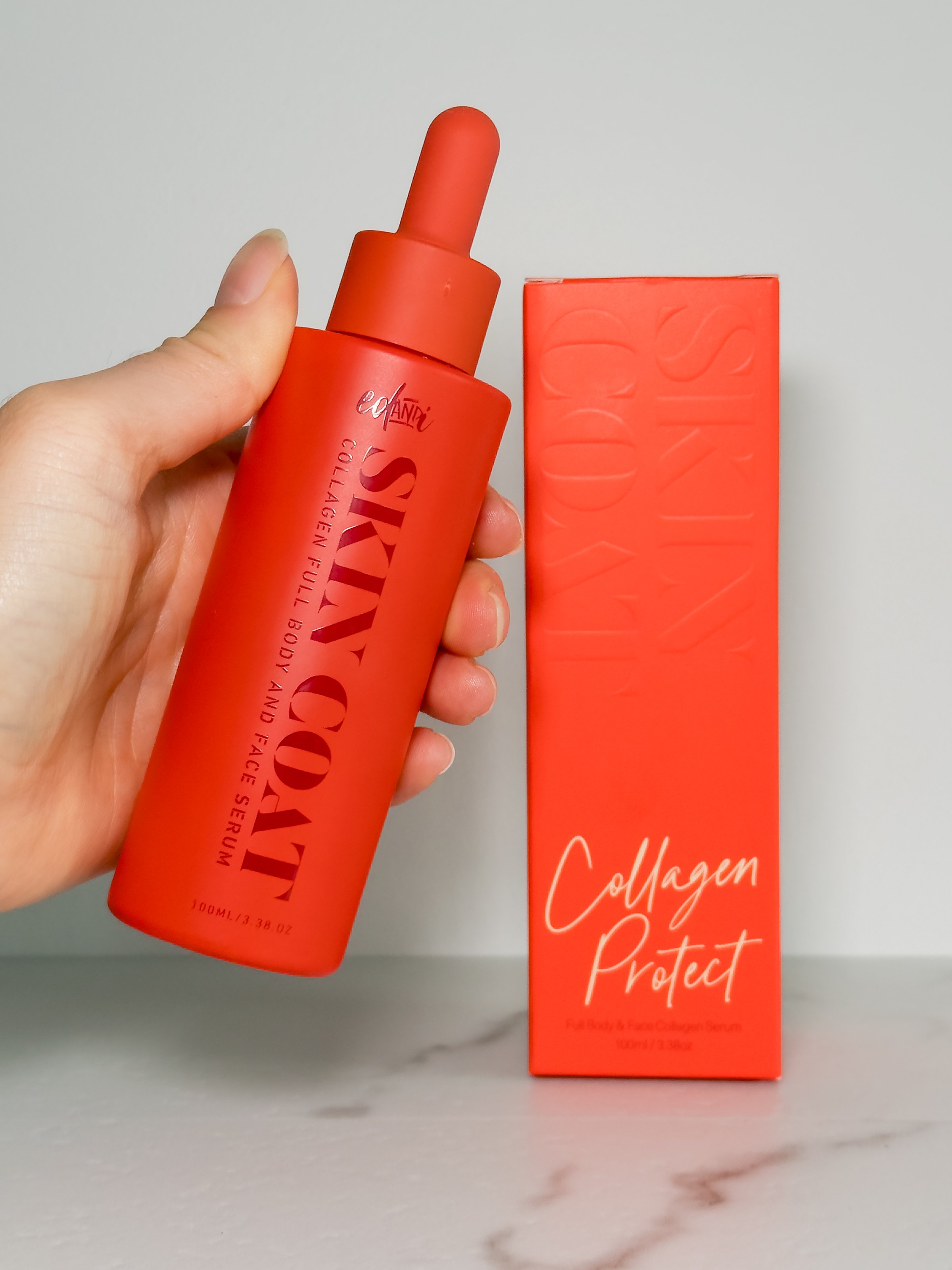 A hand holding a bright red serum bottle labeled 'SKIN COAT' next to its matching red box, both displayed on a marble surface.