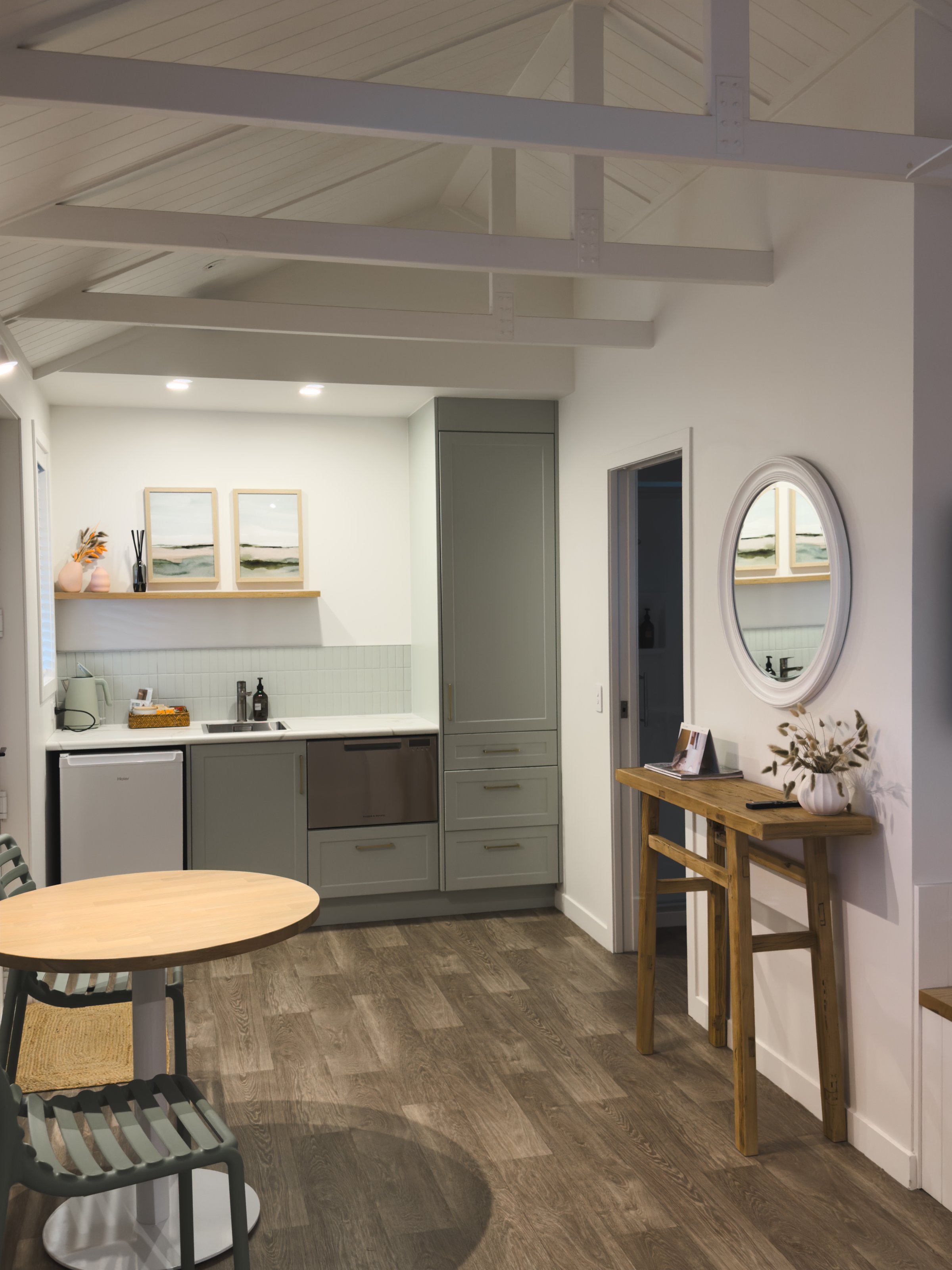 A modern kitchen with light-colored cabinetry and a round mirror on the wall. A wooden table and chairs are positioned in the foreground.