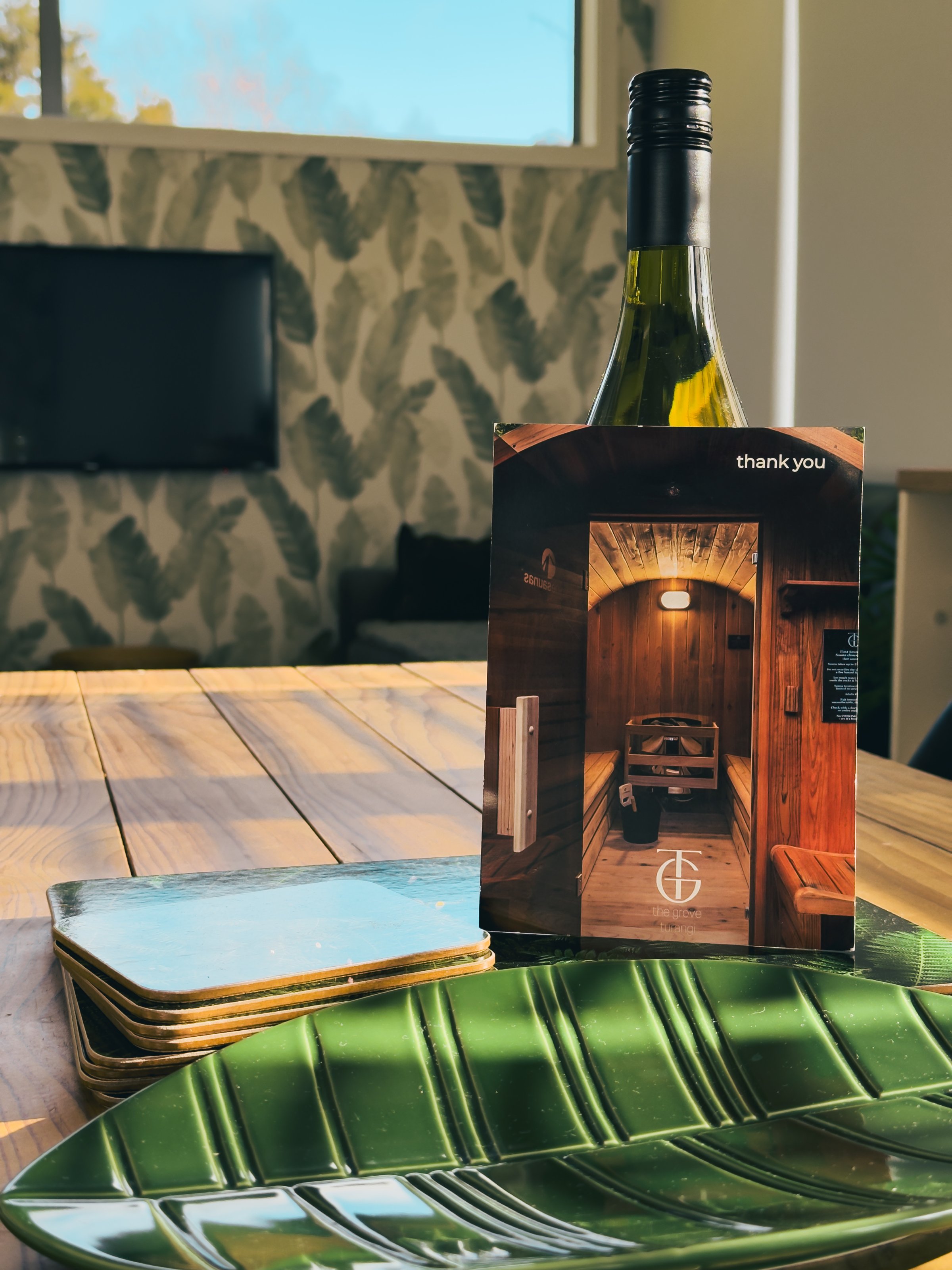 A bottle of white wine is placed on a wooden table alongside a thank you card featuring a sauna interior. A green plate and coasters are also visible in the foreground.