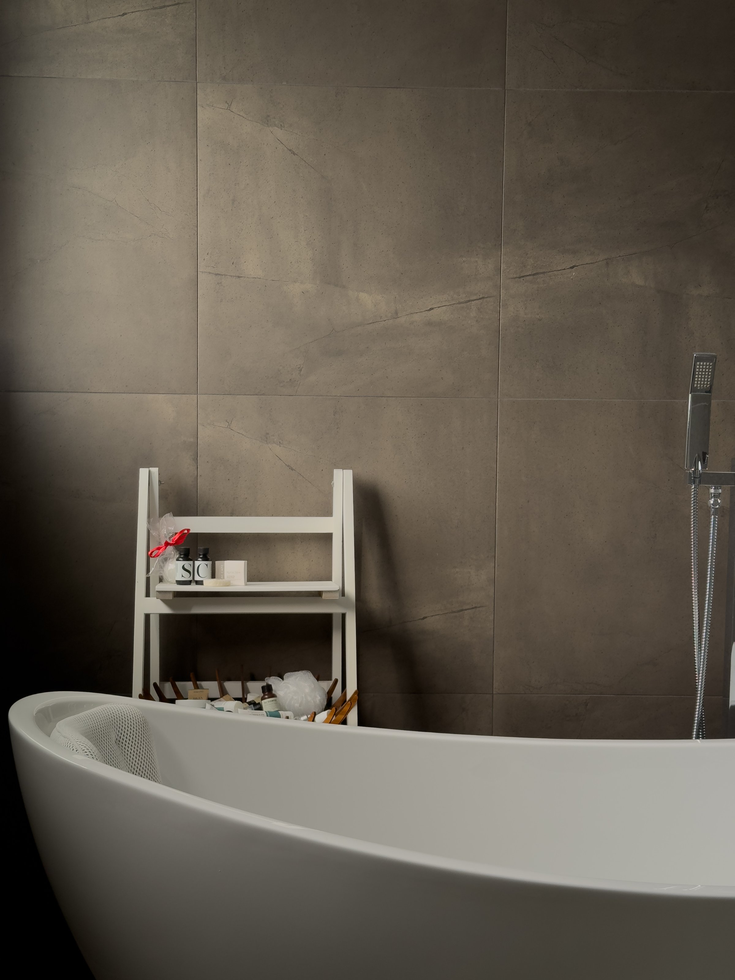 A modern white bathtub is positioned against a textured dark wall, complemented by a small white shelf holding various toiletries and a cup.