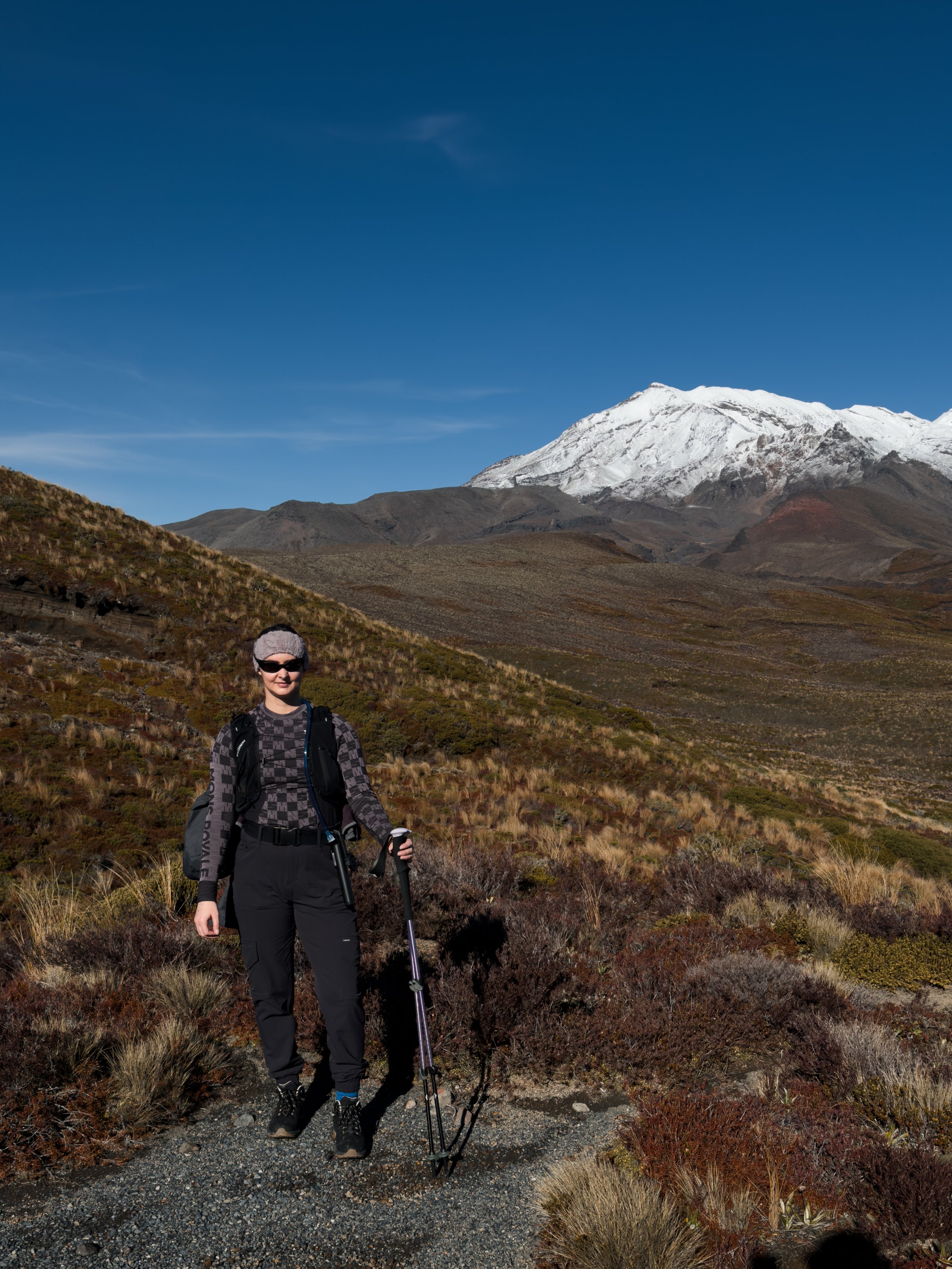 A person stands on a hiking trail with a mountainous landscape in the background, featuring snow-capped peaks and clear blue skies.
