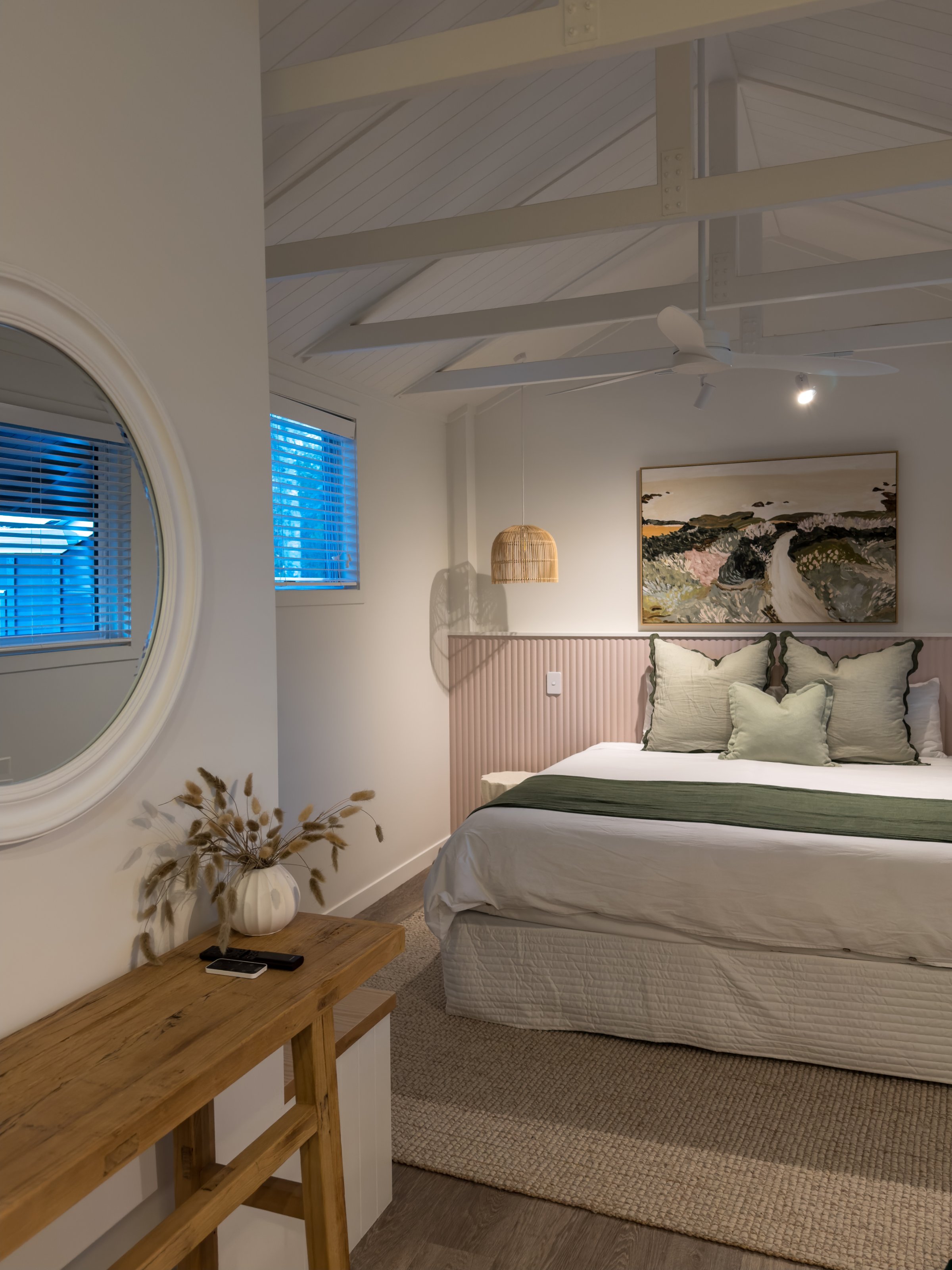 A cozy bedroom featuring a neatly made bed with green and white bedding, a round mirror on the wall, and soft natural light coming through the windows.