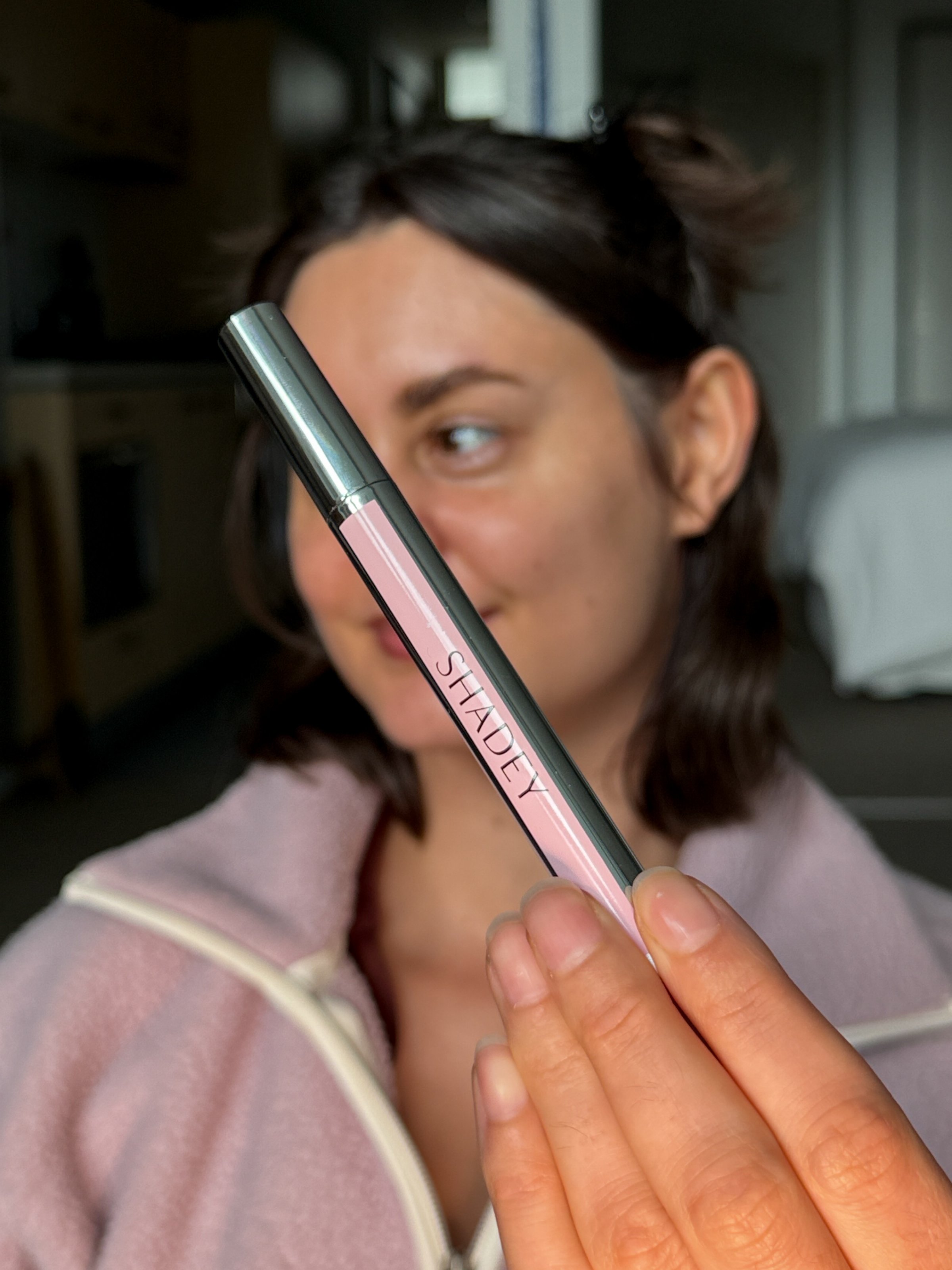 A person holding a pink cosmetic product labeled 'SHADEY' while smiling softly in a softly lit indoor setting.