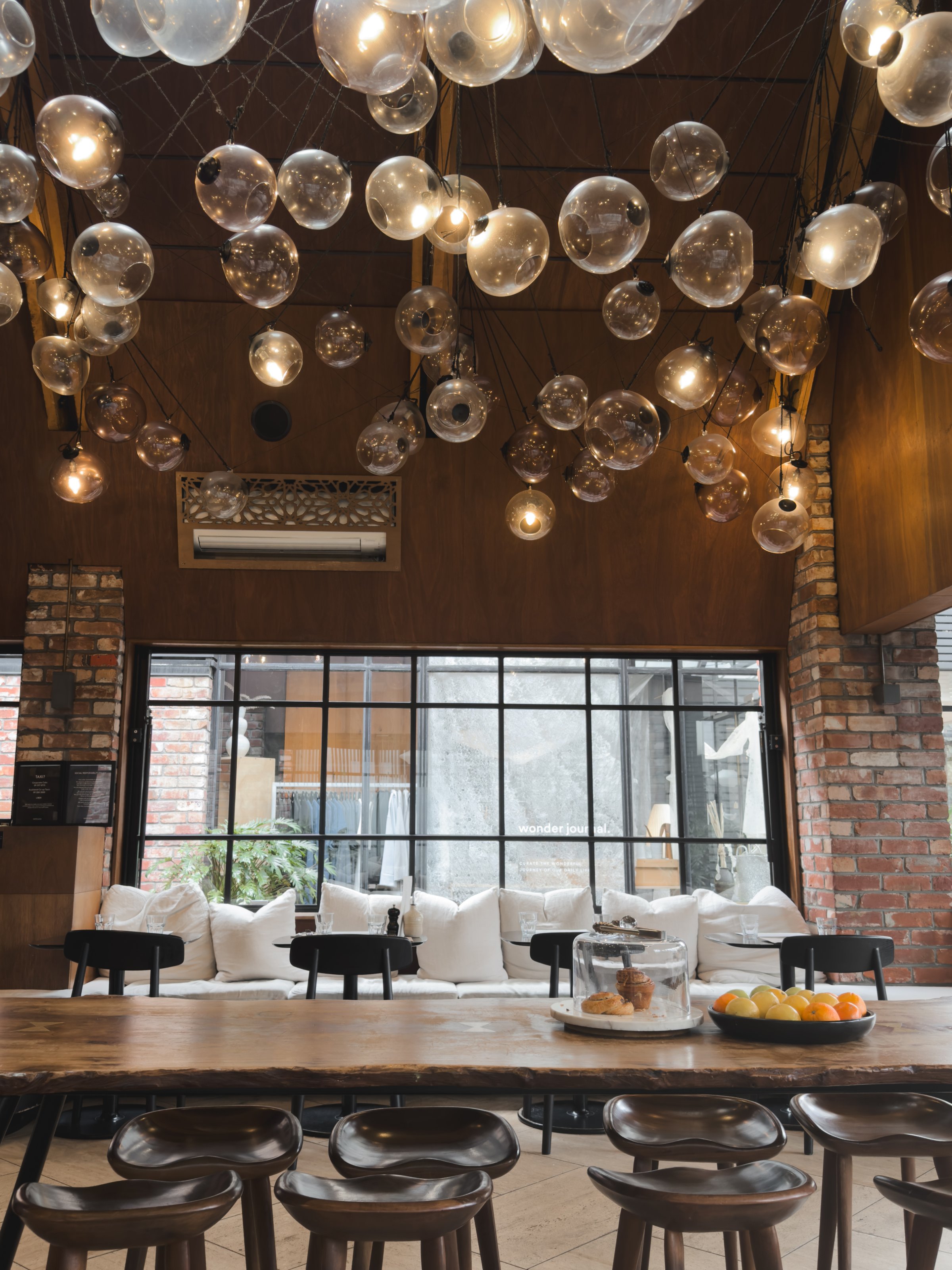 A cozy interior of a café featuring a long wooden table surrounded by dark chairs, with a backdrop of large windows and soft white cushions. The ceiling is adorned with numerous decorative glass light fixtures.