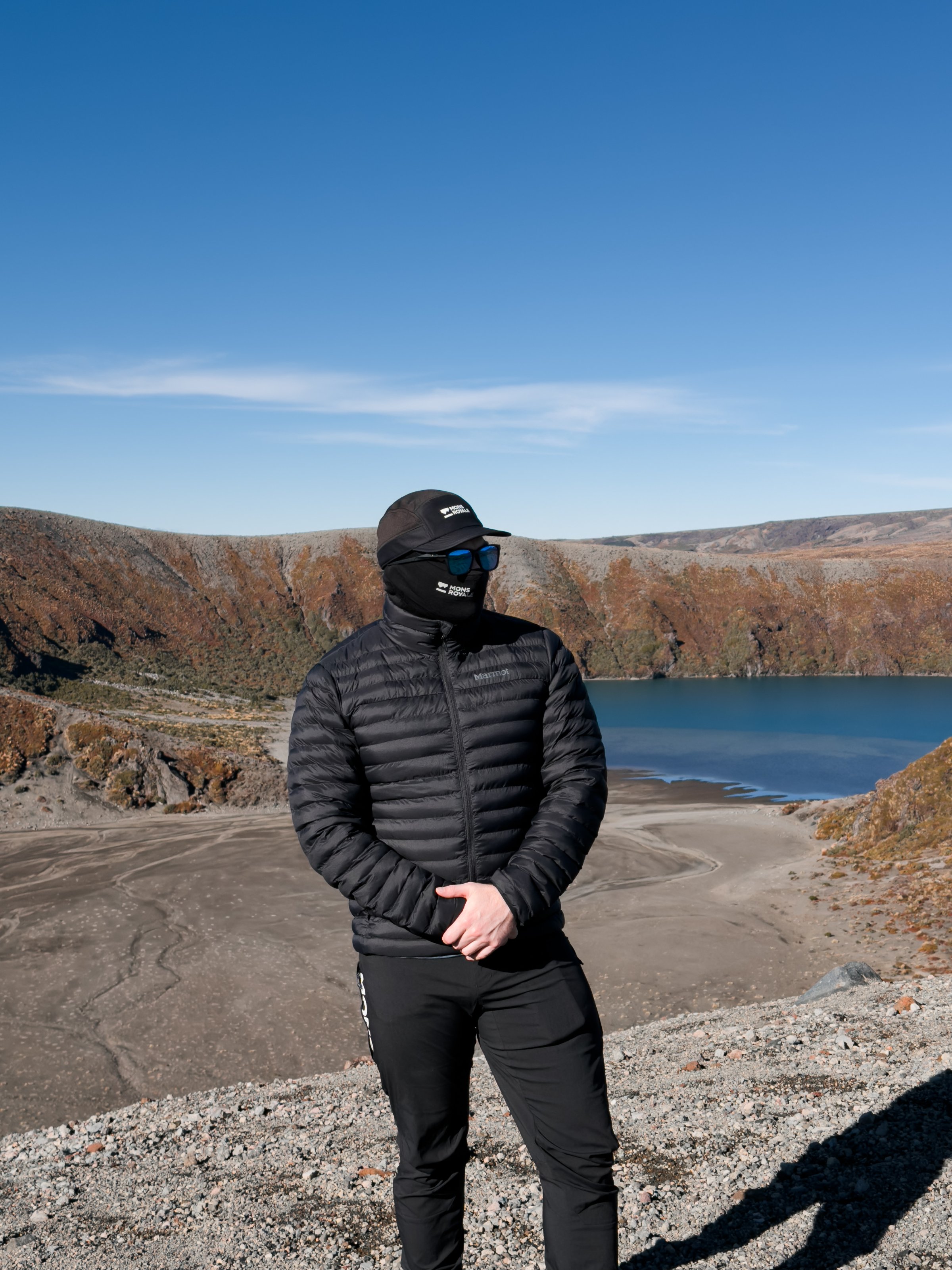 A person wearing a black puffer jacket and a cap stands near a scenic lake surrounded by rocky terrain under a clear blue sky.
