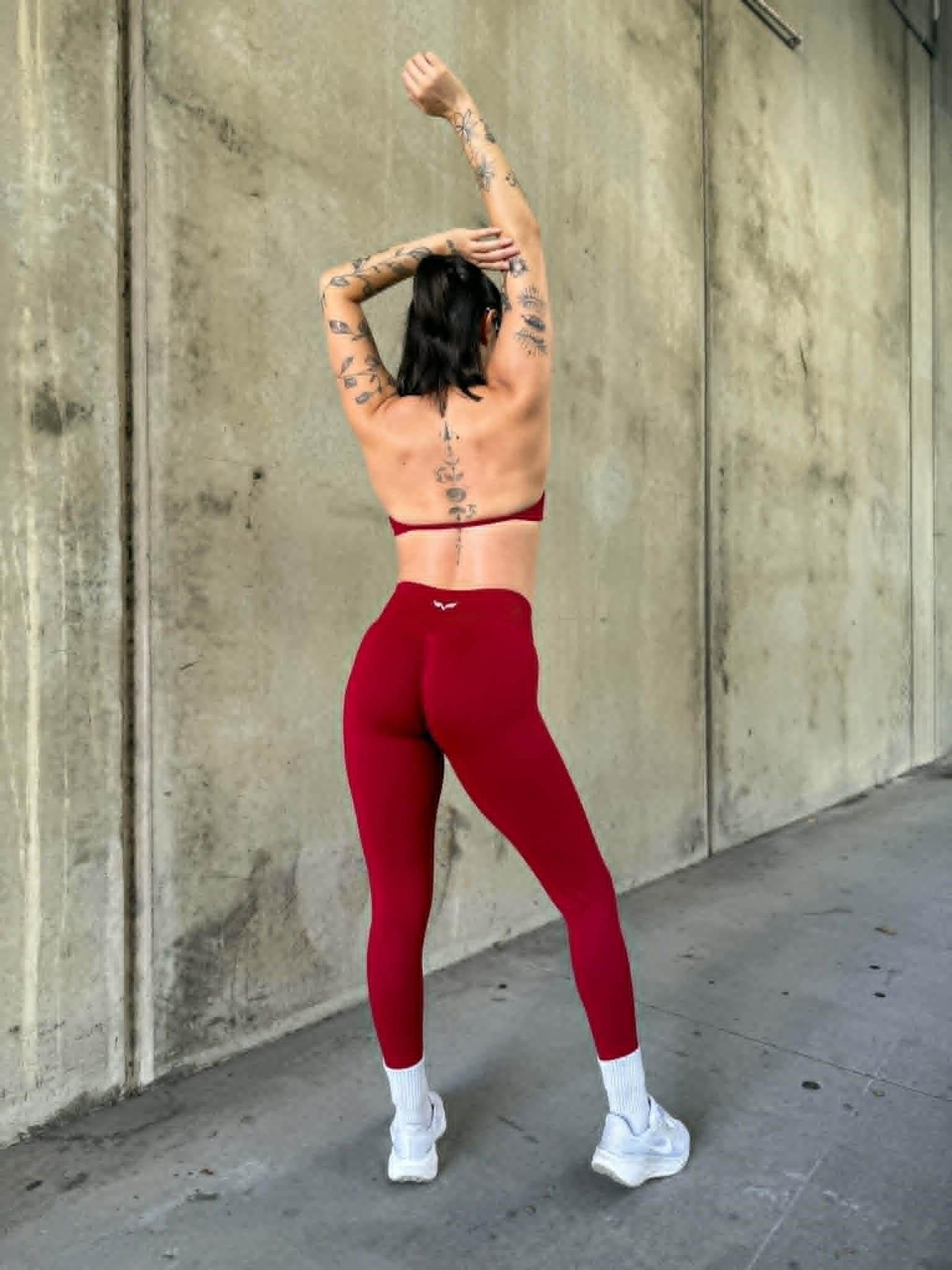 A person with tattoos poses from behind, wearing a red sports bra and matching leggings, showcasing a fitness aesthetic against a concrete backdrop.