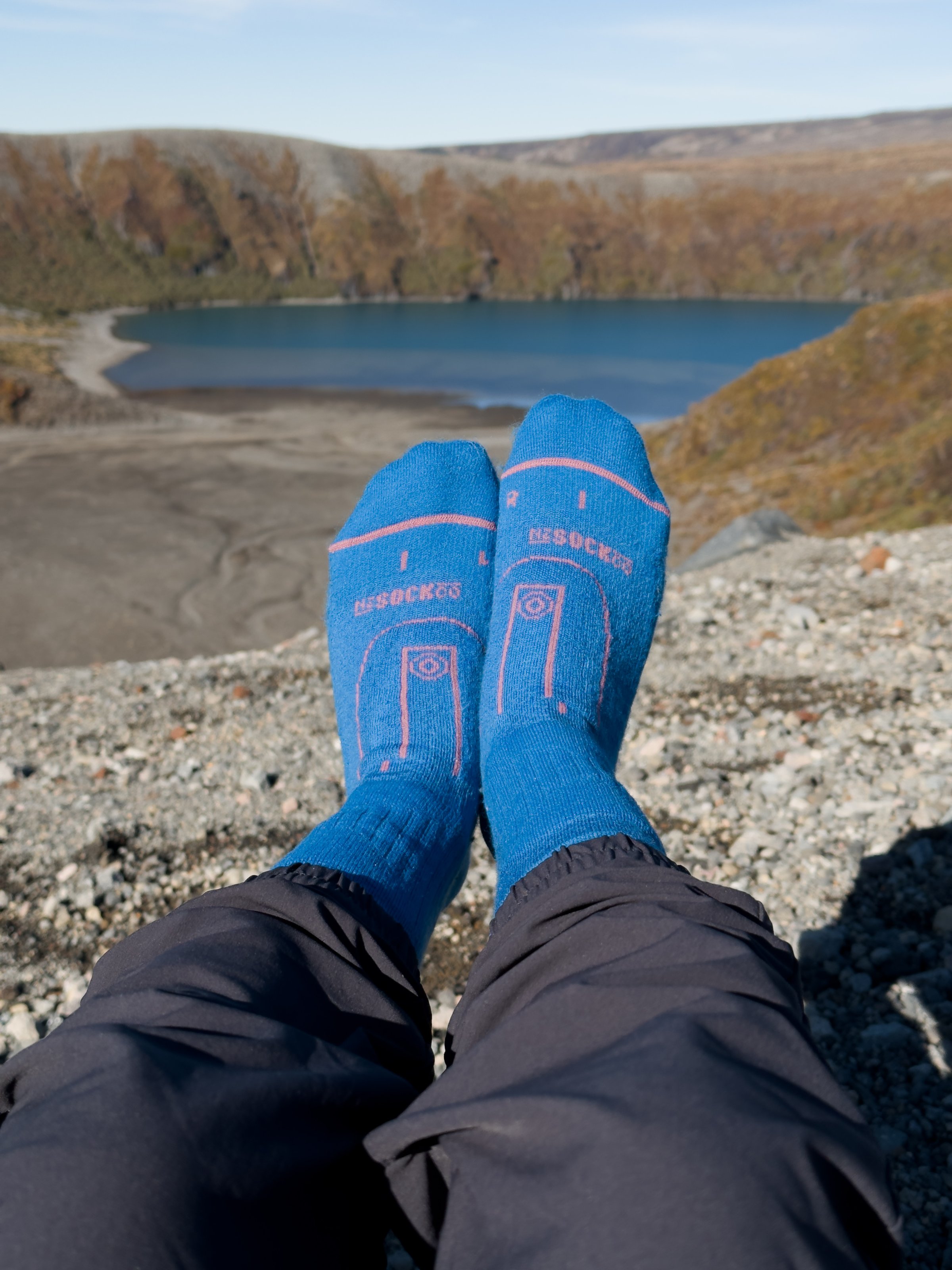 A close-up of blue socks with a graphic design, worn by a person sitting on rocky terrain overlooking a scenic lake surrounded by autumn-colored hills.