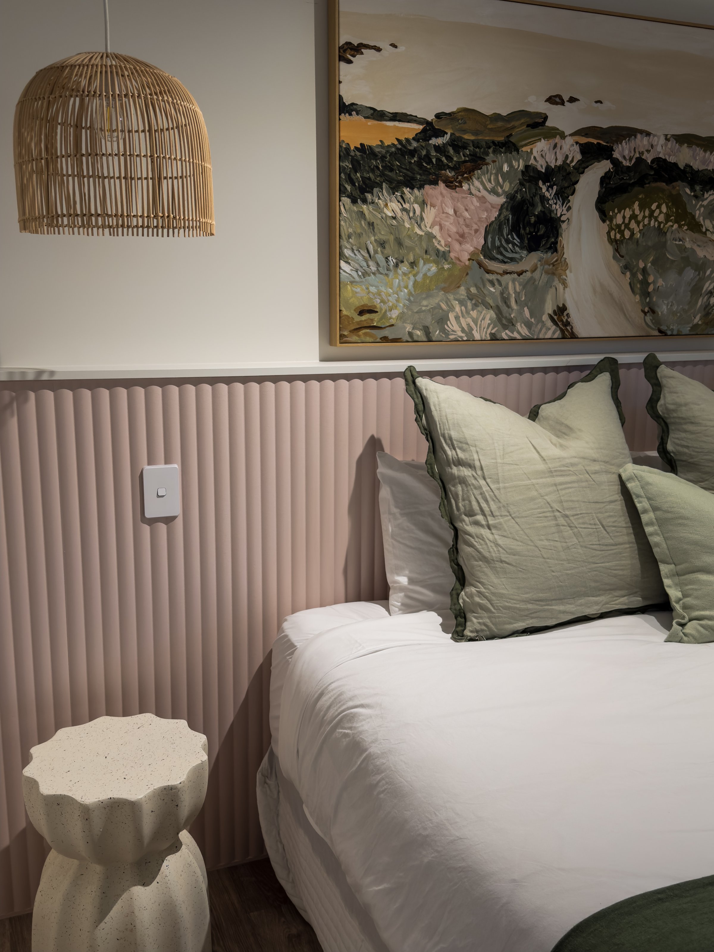 A cozy bedroom featuring a bed with green accent pillows, a textured wall, and a decorative lamp hanging above a unique side table.