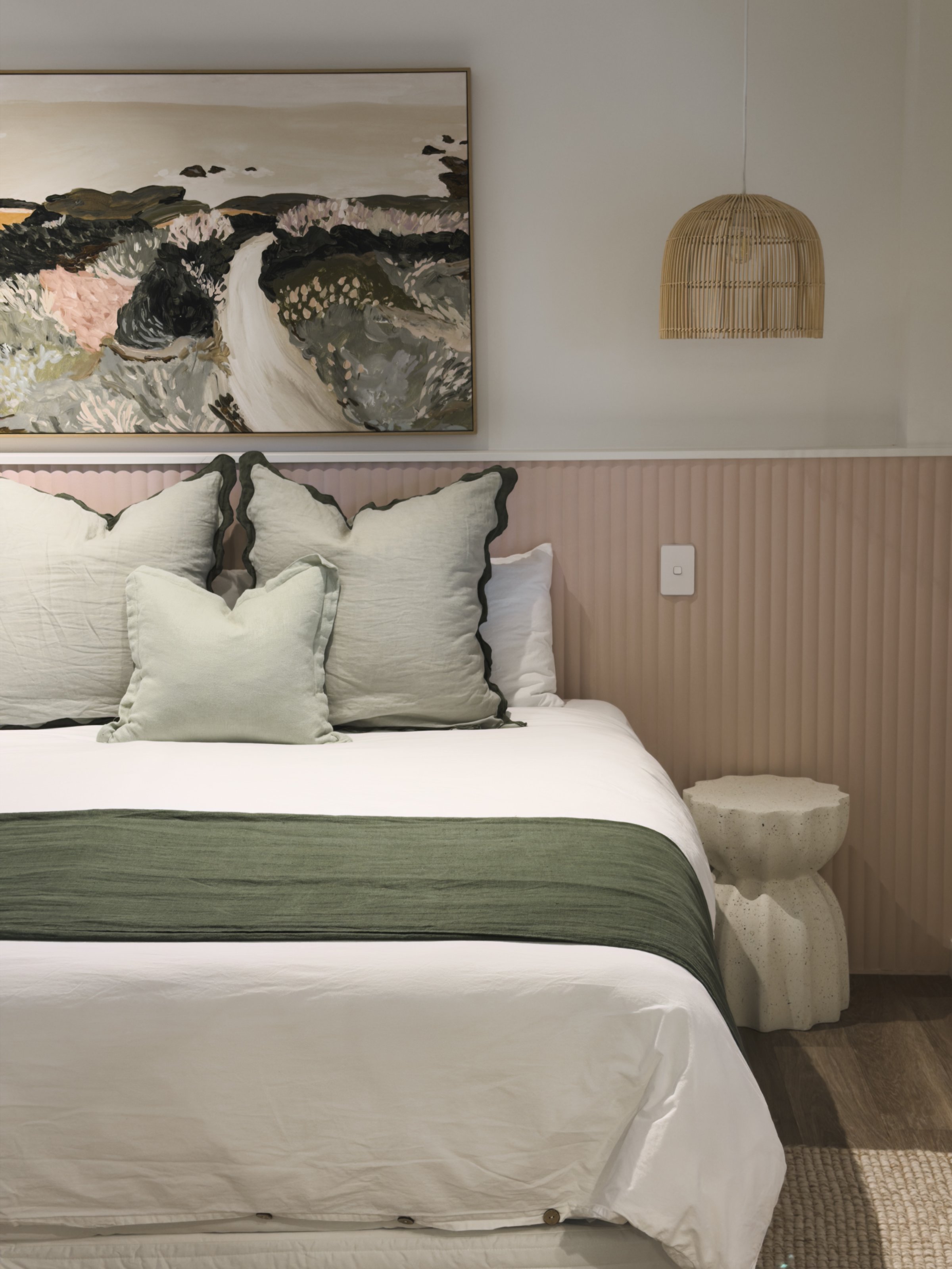 A beautifully styled bedroom featuring a white bed with green accents, decorative pillows, and a piece of artwork above the headboard. A woven pendant light hangs from the ceiling, adding a cozy touch.