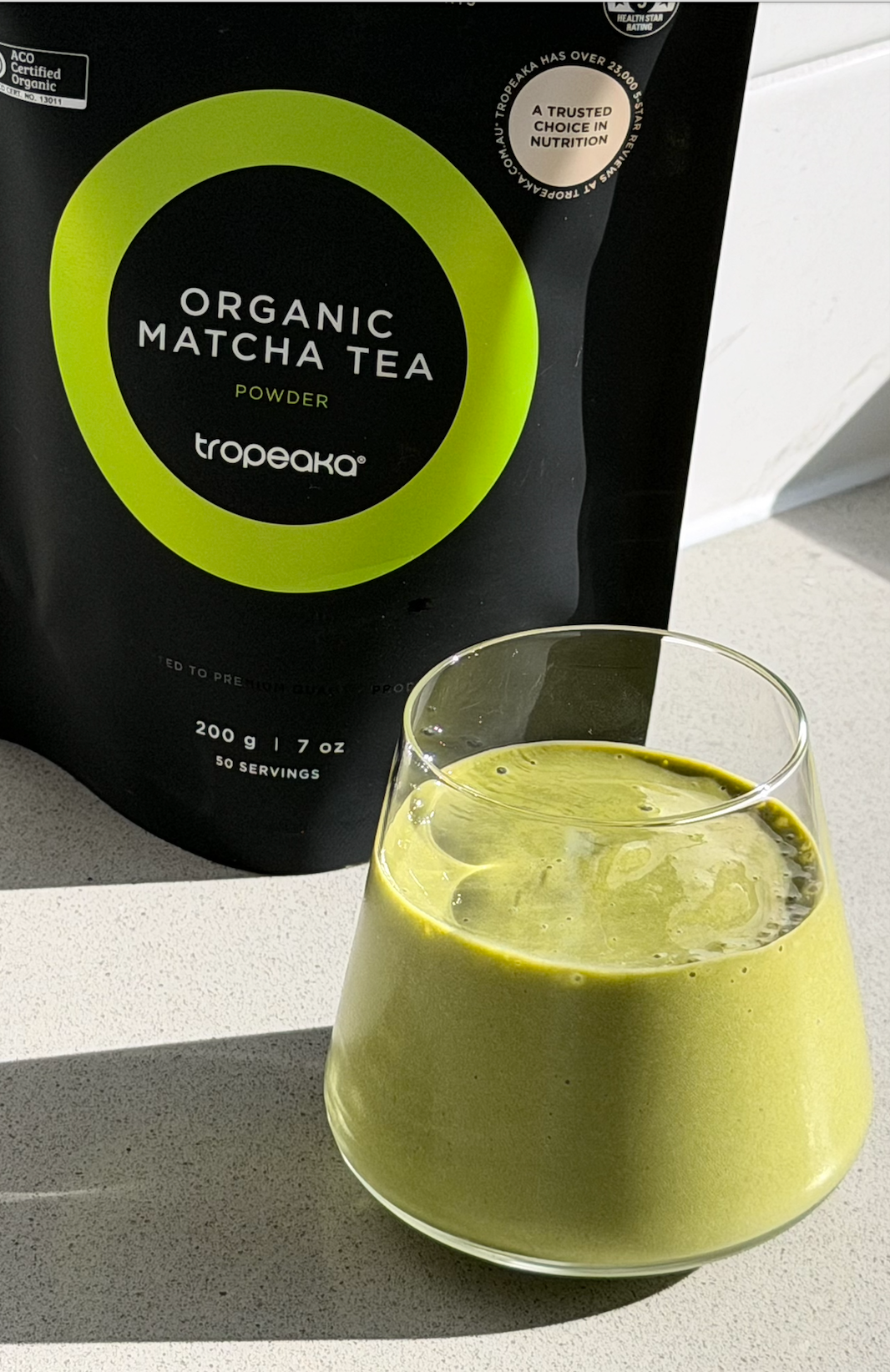 A glass of vibrant green matcha tea sits on a light-colored countertop next to a black package of organic matcha tea powder.