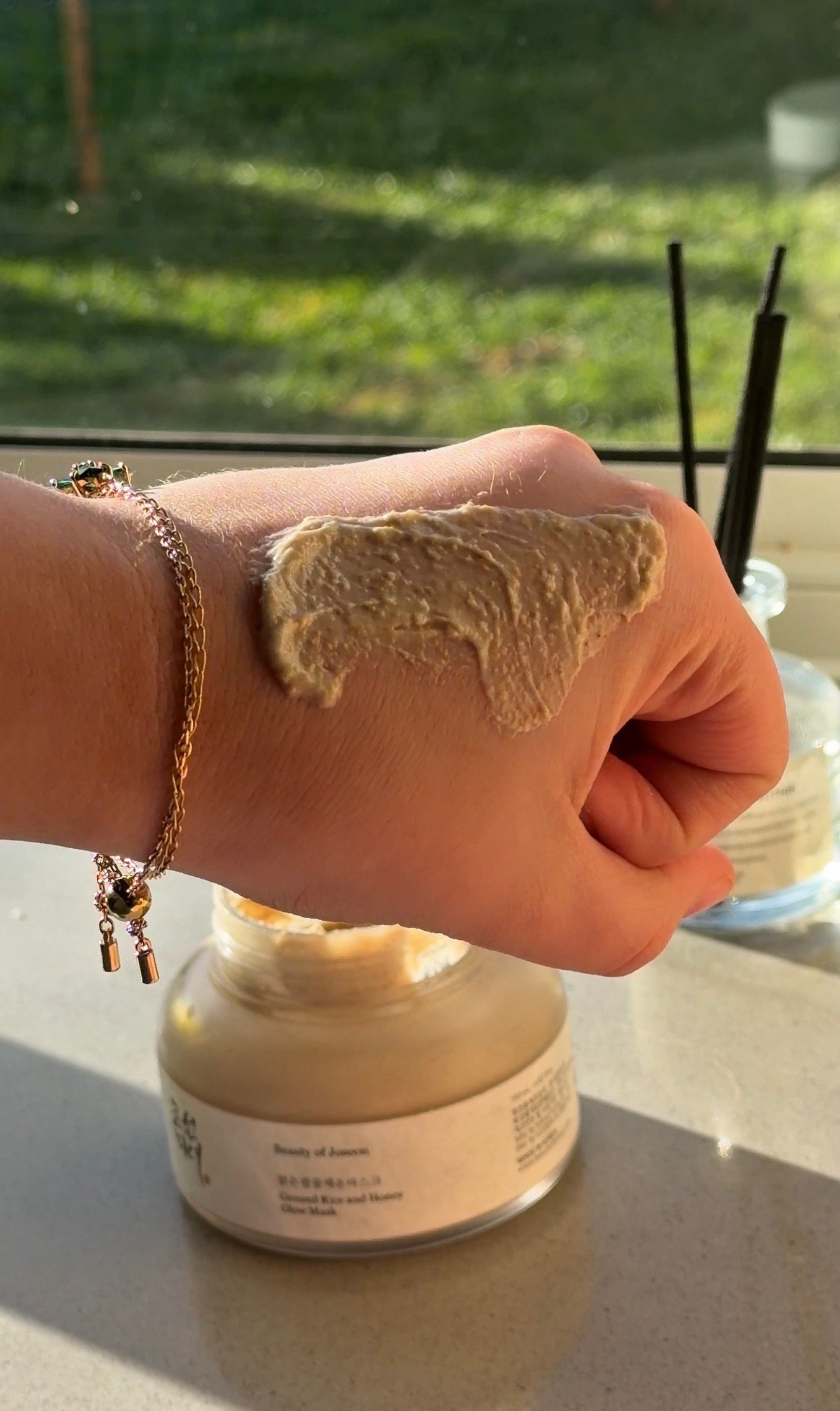 A hand with a creamy product applied to the skin, placed next to a jar of skincare on a countertop with natural light streaming in.