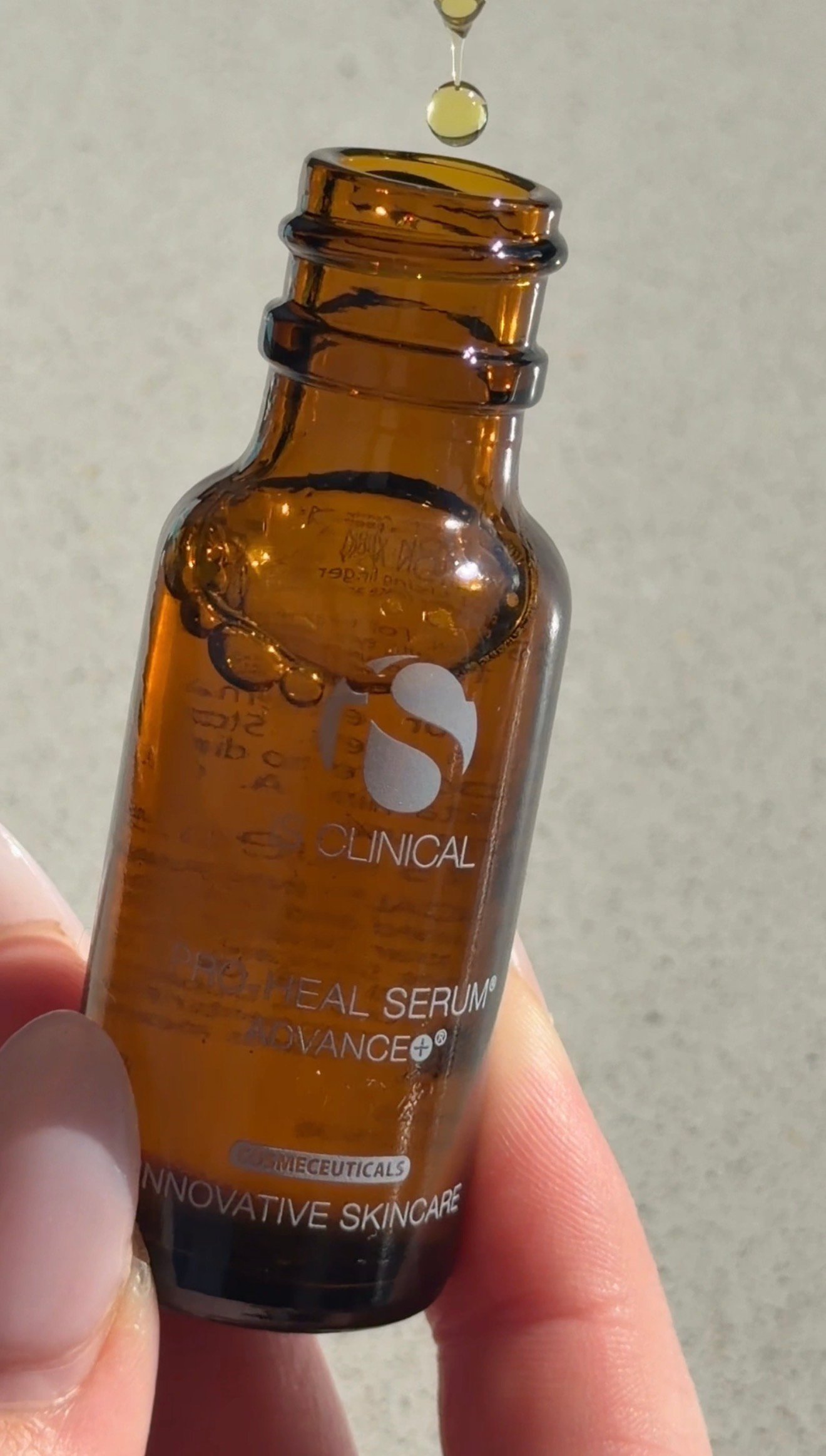 A close-up of a hand holding a small amber glass bottle of skincare serum, with a drop of serum poised above the opening.