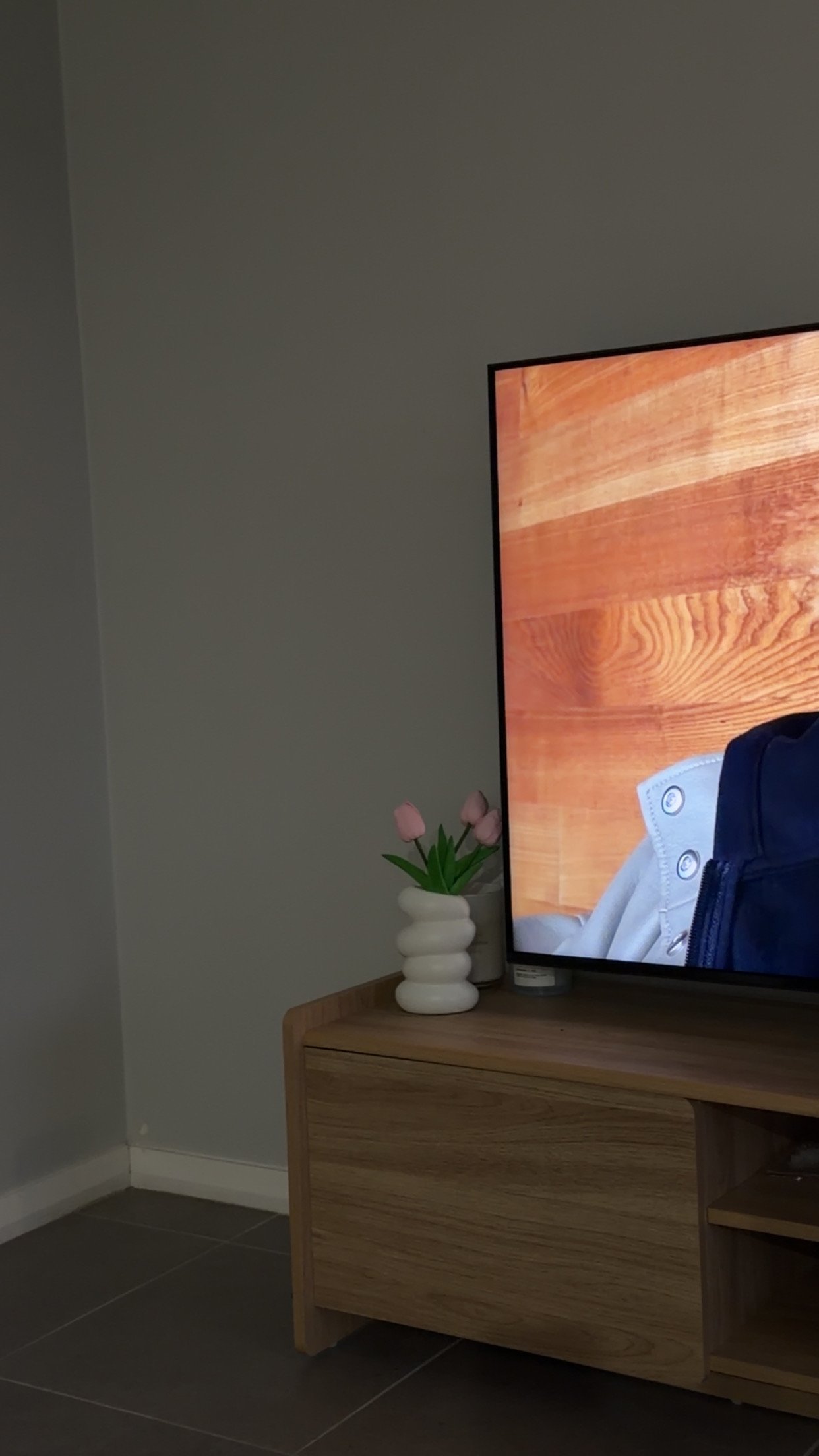 A cozy living room scene featuring a television displaying a person, with a wooden wall in the background and a decorative vase of tulips on a wooden cabinet.