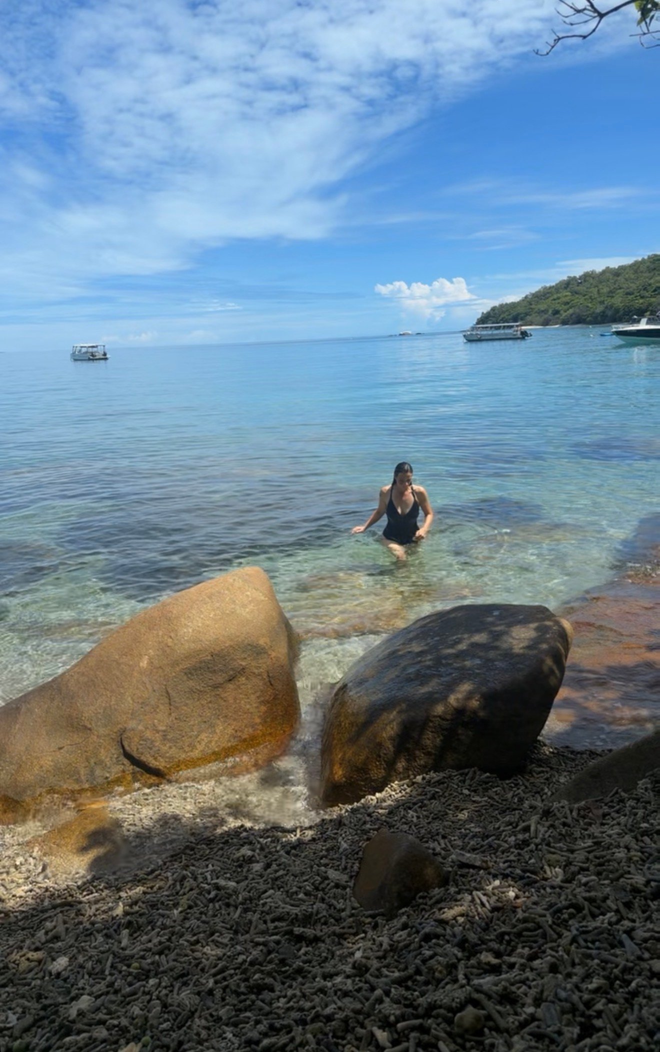 A person wading in clear water near a rocky shore, surrounded by lush greenery and boats in the distance under a blue sky.