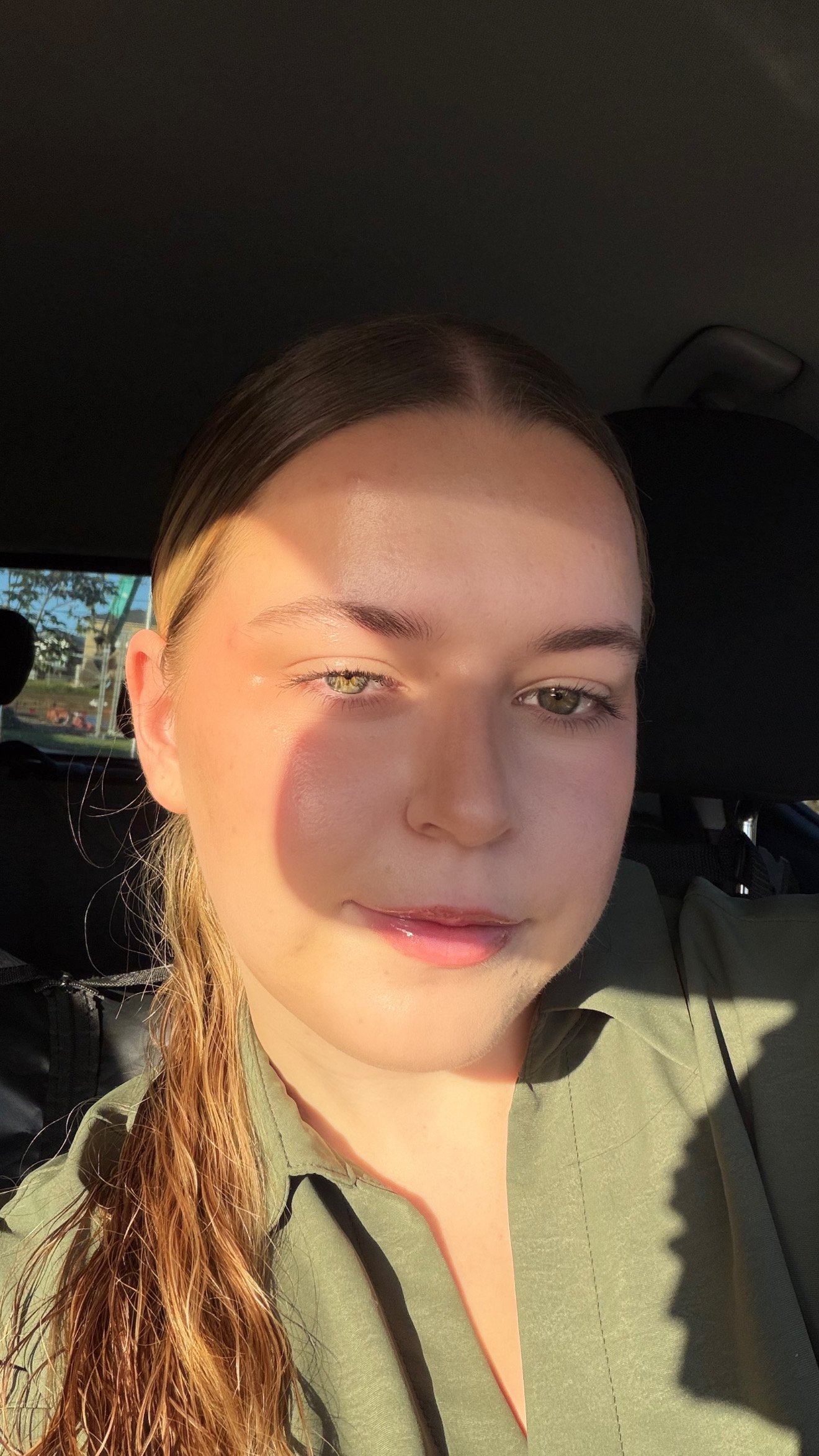 A close-up portrait of a young woman with long hair, sitting in a car. Soft sunlight highlights her face, creating a warm glow.