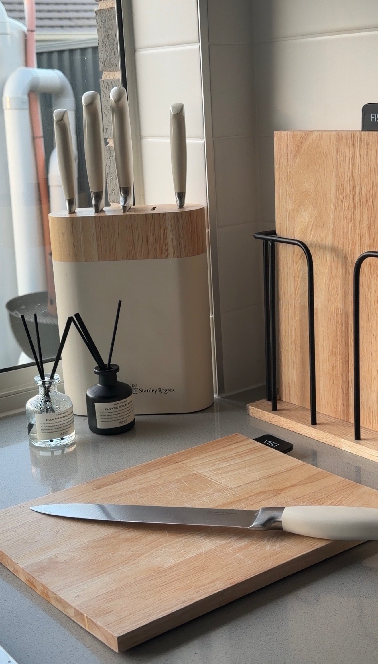 A modern kitchen countertop featuring a wooden cutting board, a sleek knife, and a knife block with several knives. Decorative items like a diffuser and a candle add a stylish touch.