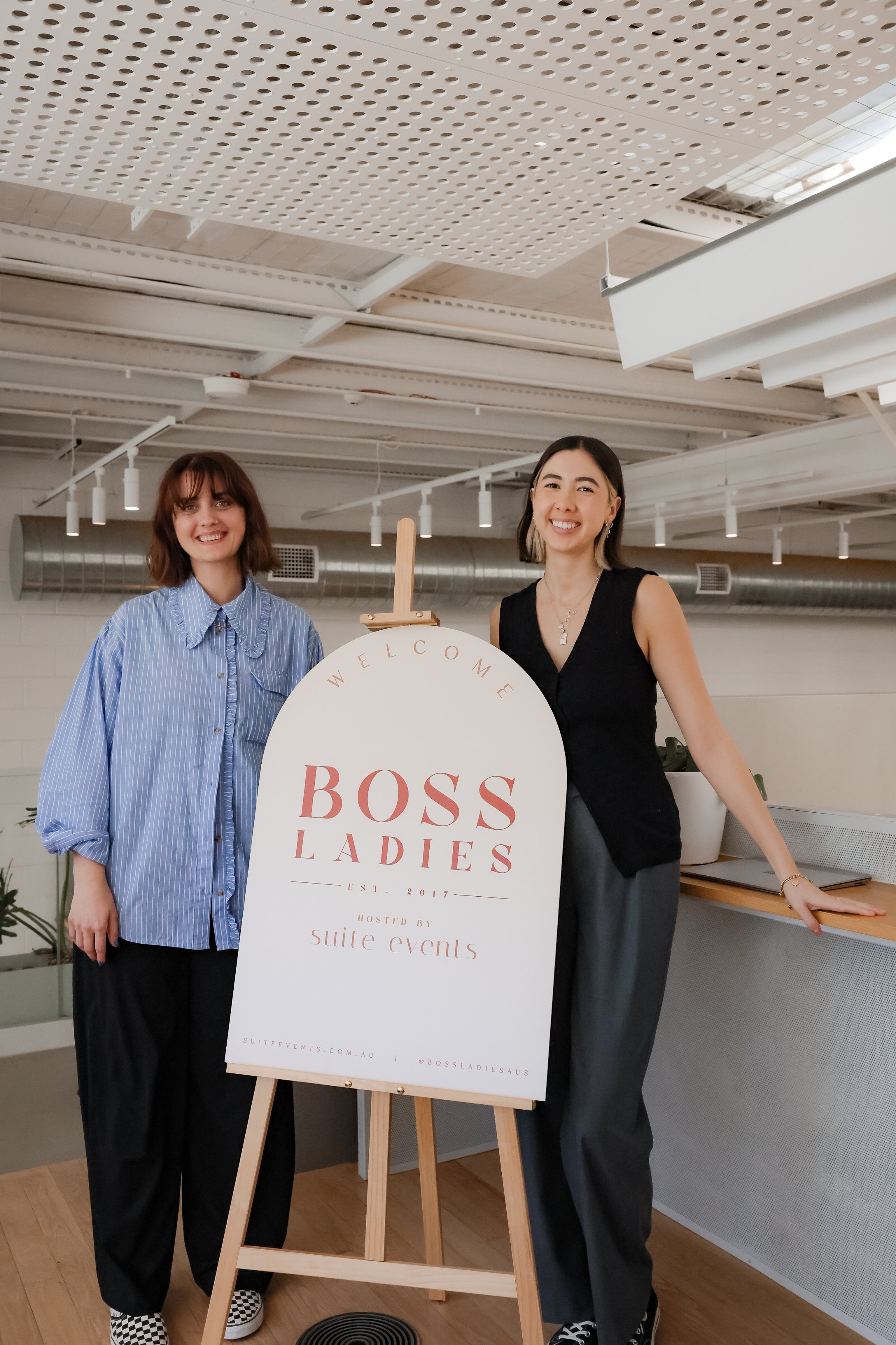 Two women stand beside a welcome sign for an event titled 'BOSS LADIES' in a modern indoor space.
