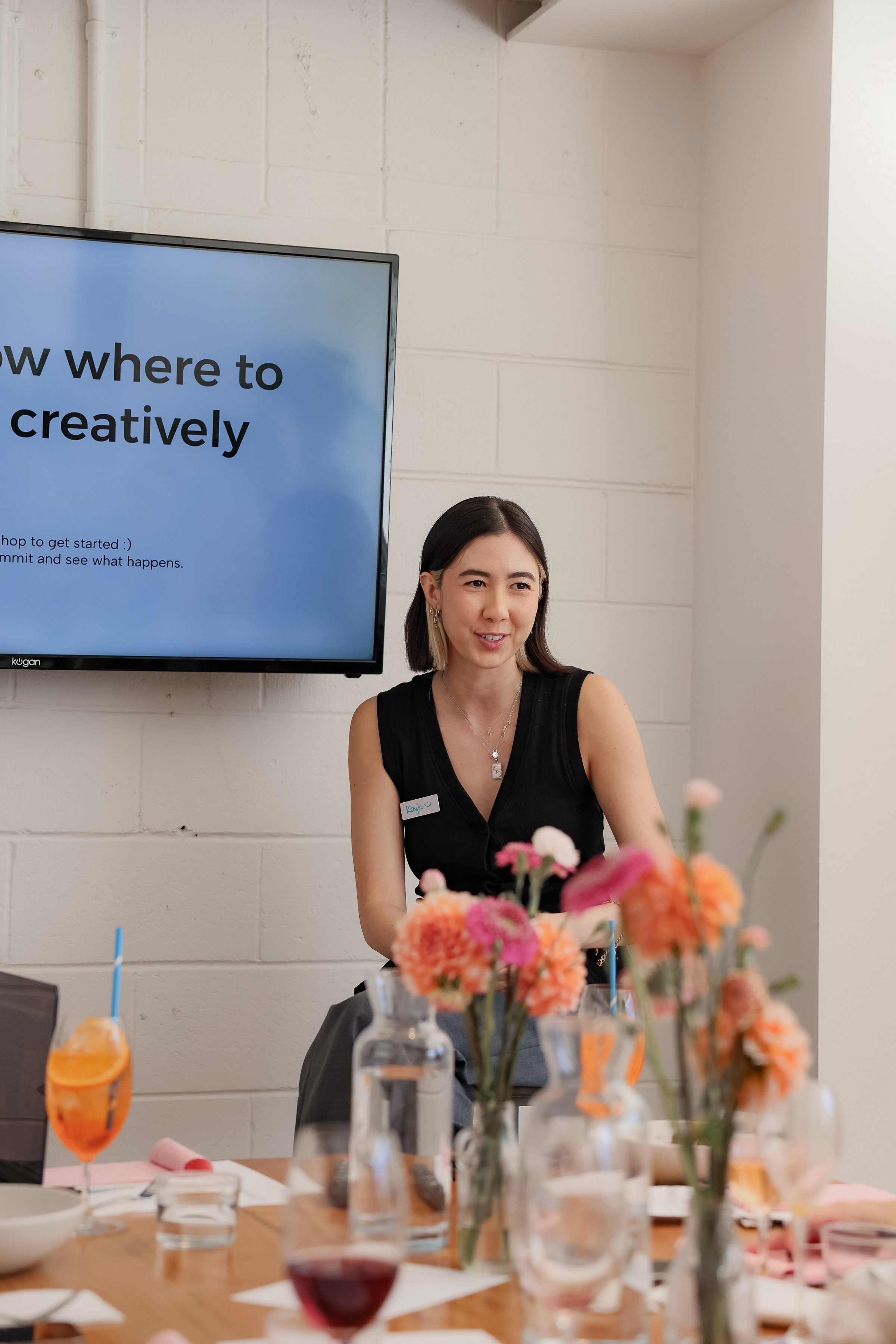 A woman in a black top speaks at a workshop, with a presentation screen in the background and a table adorned with flowers and drinks.