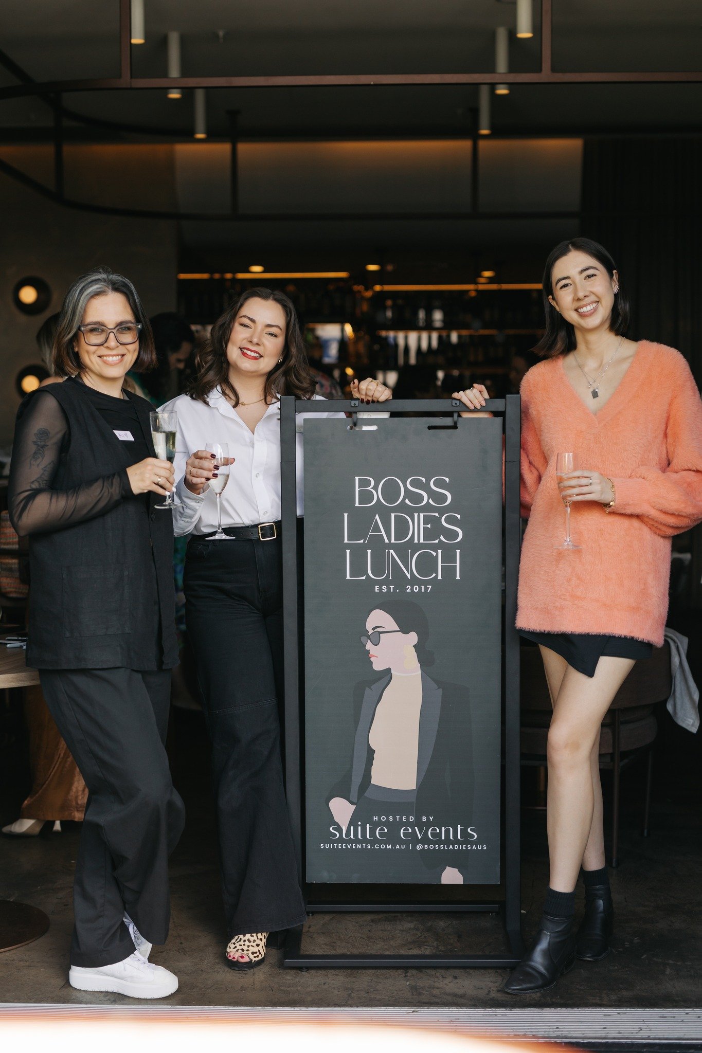 Three women pose together at a social event, holding drinks in front of a sign that reads 'BOSS LADIES LUNCH'. The setting is stylish and modern.