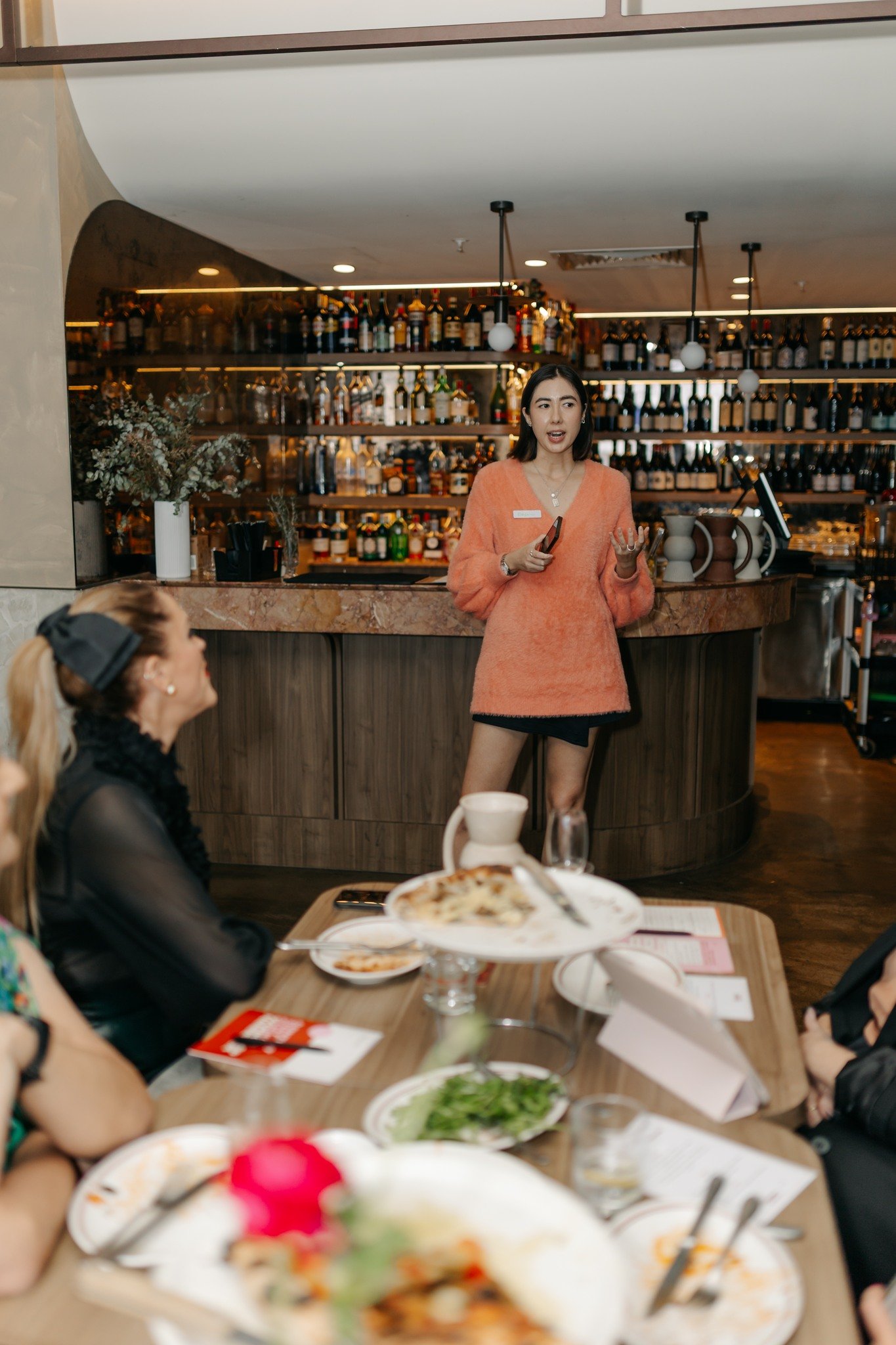 A woman in an orange sweater stands and speaks at a restaurant, with a bar filled with bottles in the background. Several seated guests listen attentively at a table covered with food.