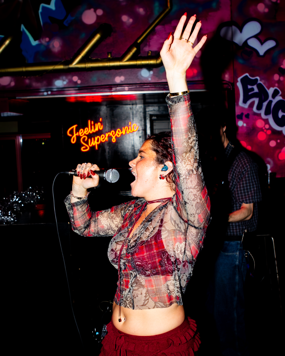 A performer passionately singing into a microphone with one hand raised, set against a vibrant, graffiti-covered backdrop.