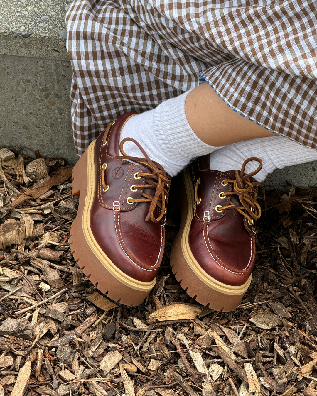 A close-up of brown platform shoes paired with white socks, resting on a bed of mulch, with a checkered skirt partially visible.