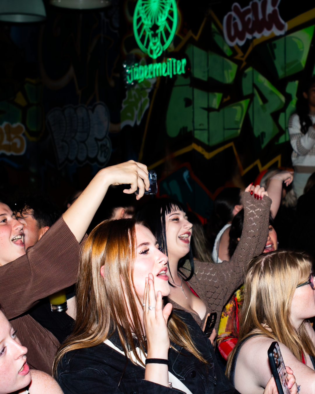 A lively crowd enjoying a performance, with people cheering and taking photos against a colorful graffiti backdrop.