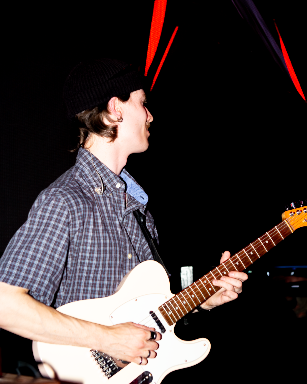 A musician playing an electric guitar on stage, captured from the side with vibrant lighting in the background.