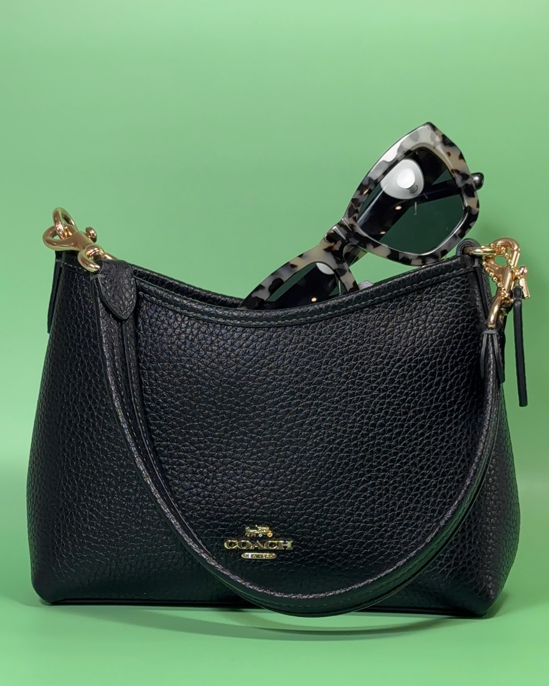 A black leather handbag with a textured finish, featuring a gold logo, alongside a pair of stylish tortoiseshell sunglasses against a green background.