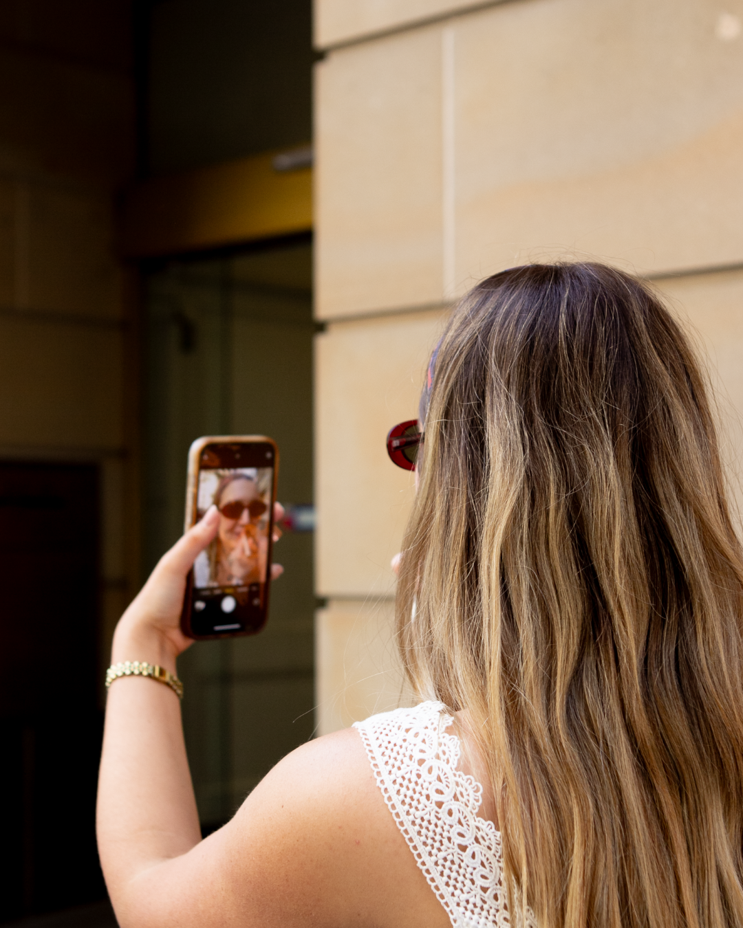 A woman with long hair takes a selfie with her phone, wearing sunglasses, in an urban setting.
