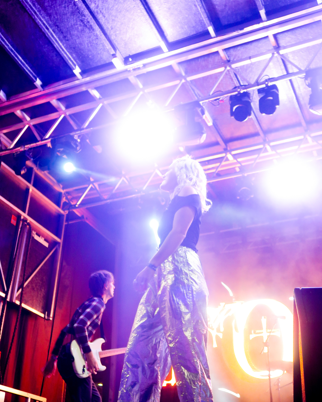 A performer on stage with bright lights and a guitarist in the background, creating an energetic concert atmosphere.