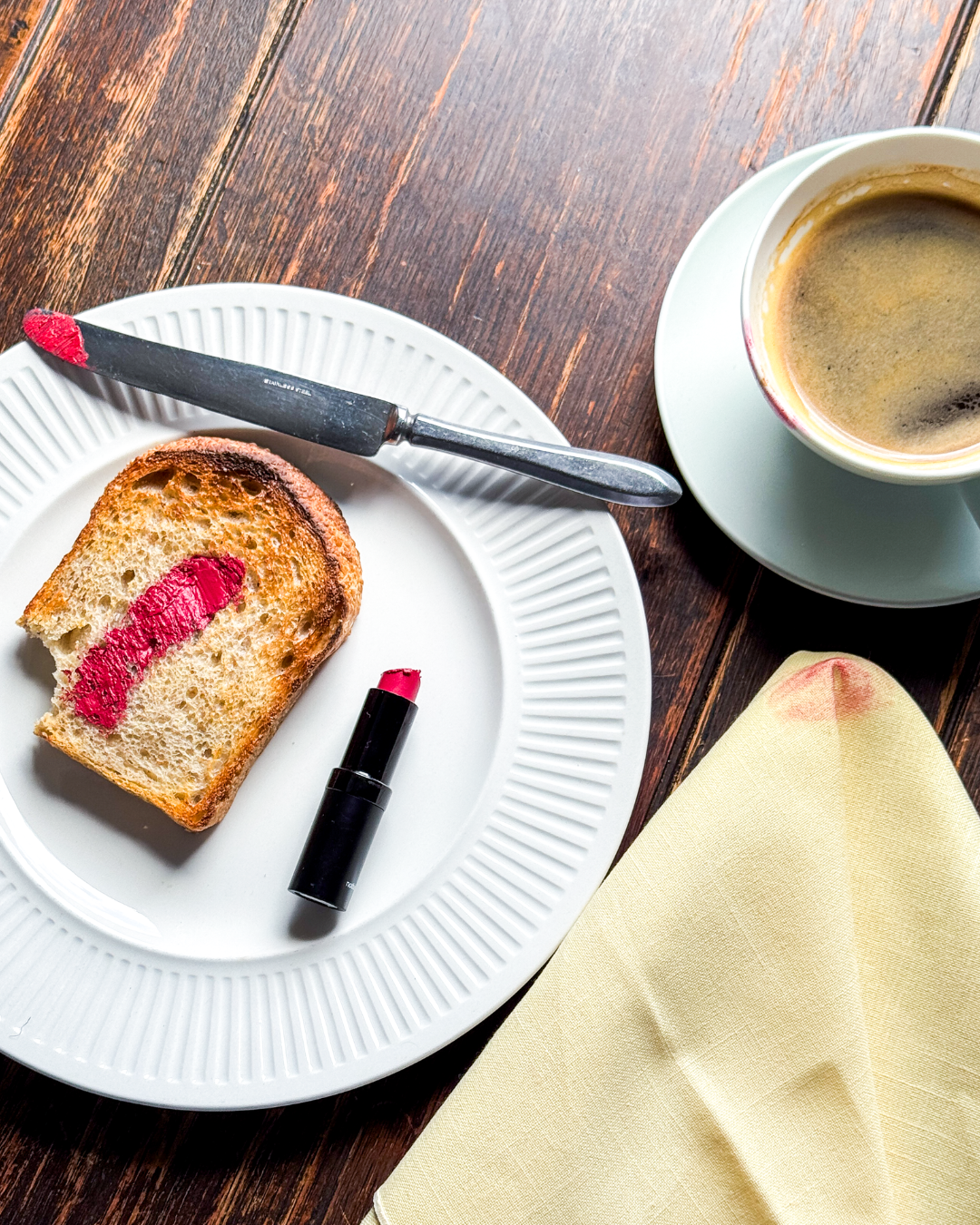 A slice of bread with a lipstick mark on it, accompanied by a coffee cup and a folded napkin on a wooden table.