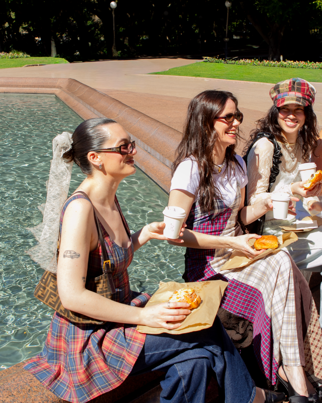 Three women enjoying pastries and coffee by a fountain in a park, dressed in stylish plaid outfits.