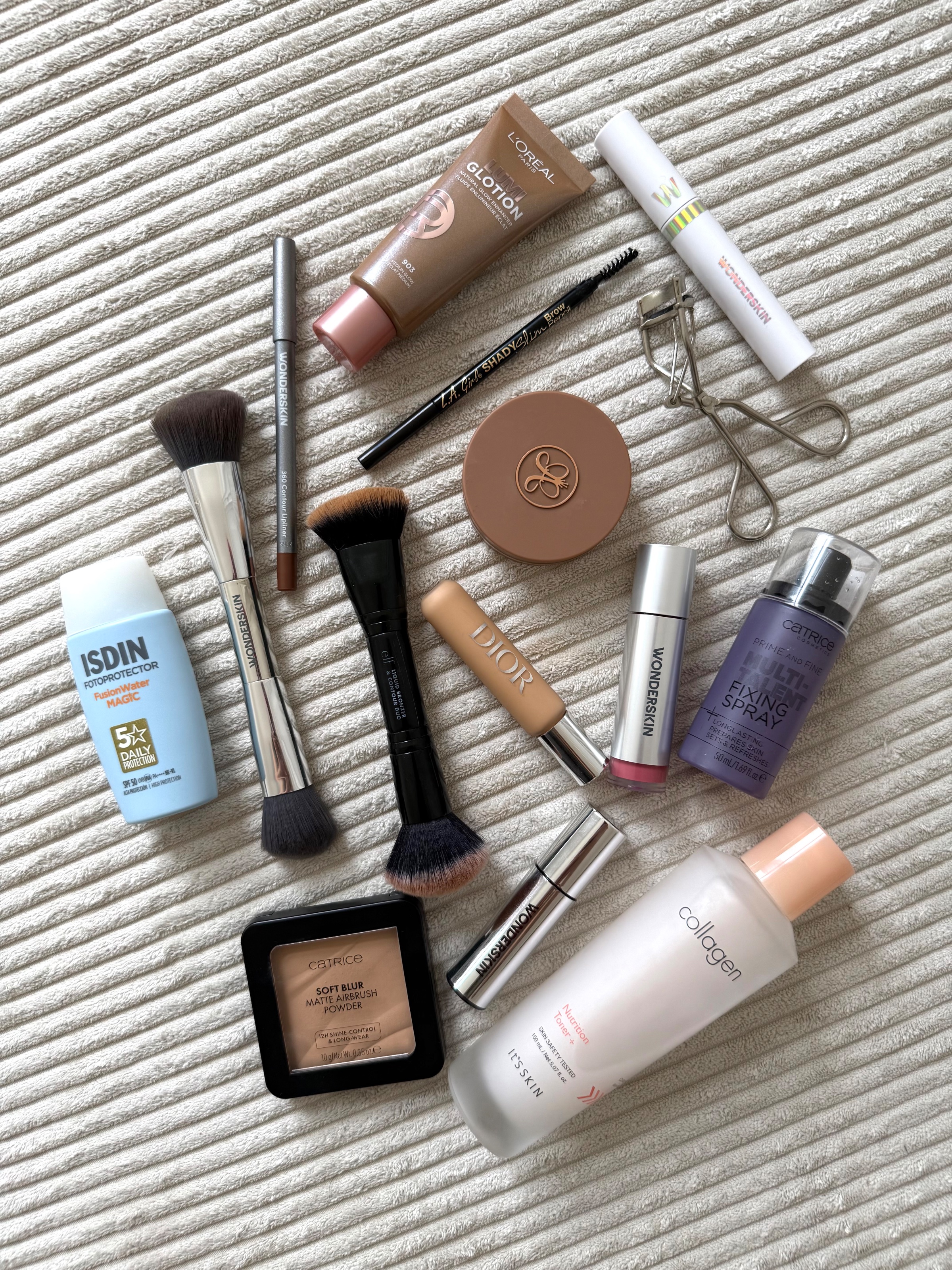 A flat lay arrangement of various makeup and skincare products on a textured surface, including foundations, brushes, and a sunscreen.