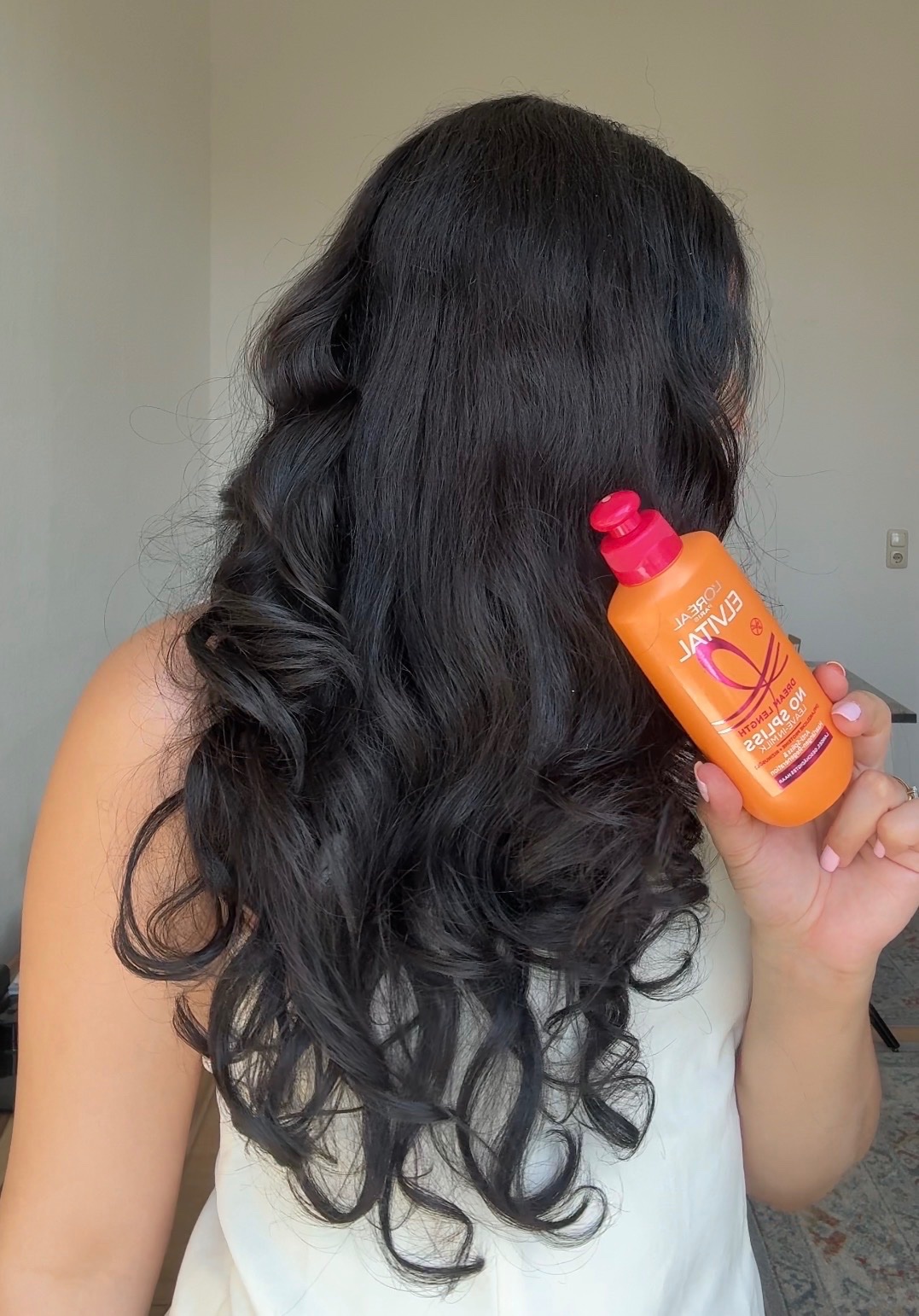 A person with long, wavy black hair holds a bottle of hair product in front of them, showcasing the product's vibrant packaging.