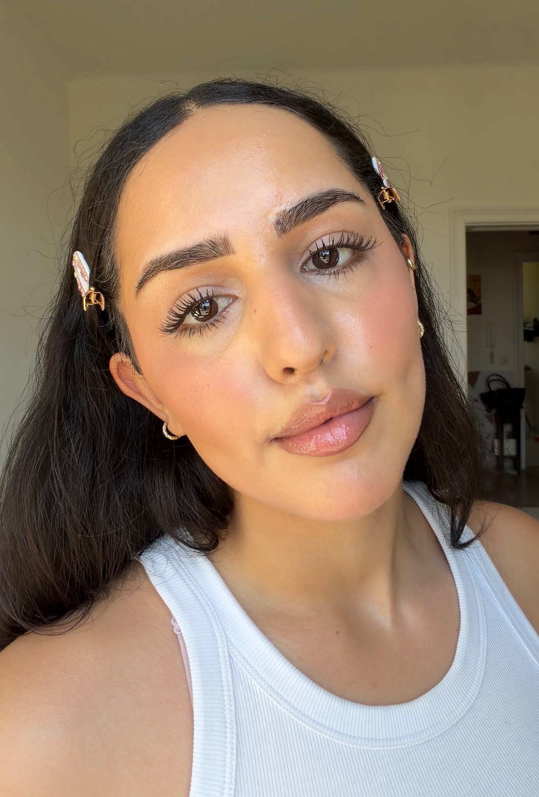 A close-up portrait of a woman with long dark hair, wearing a white tank top and hair clips, showcasing a natural makeup look.