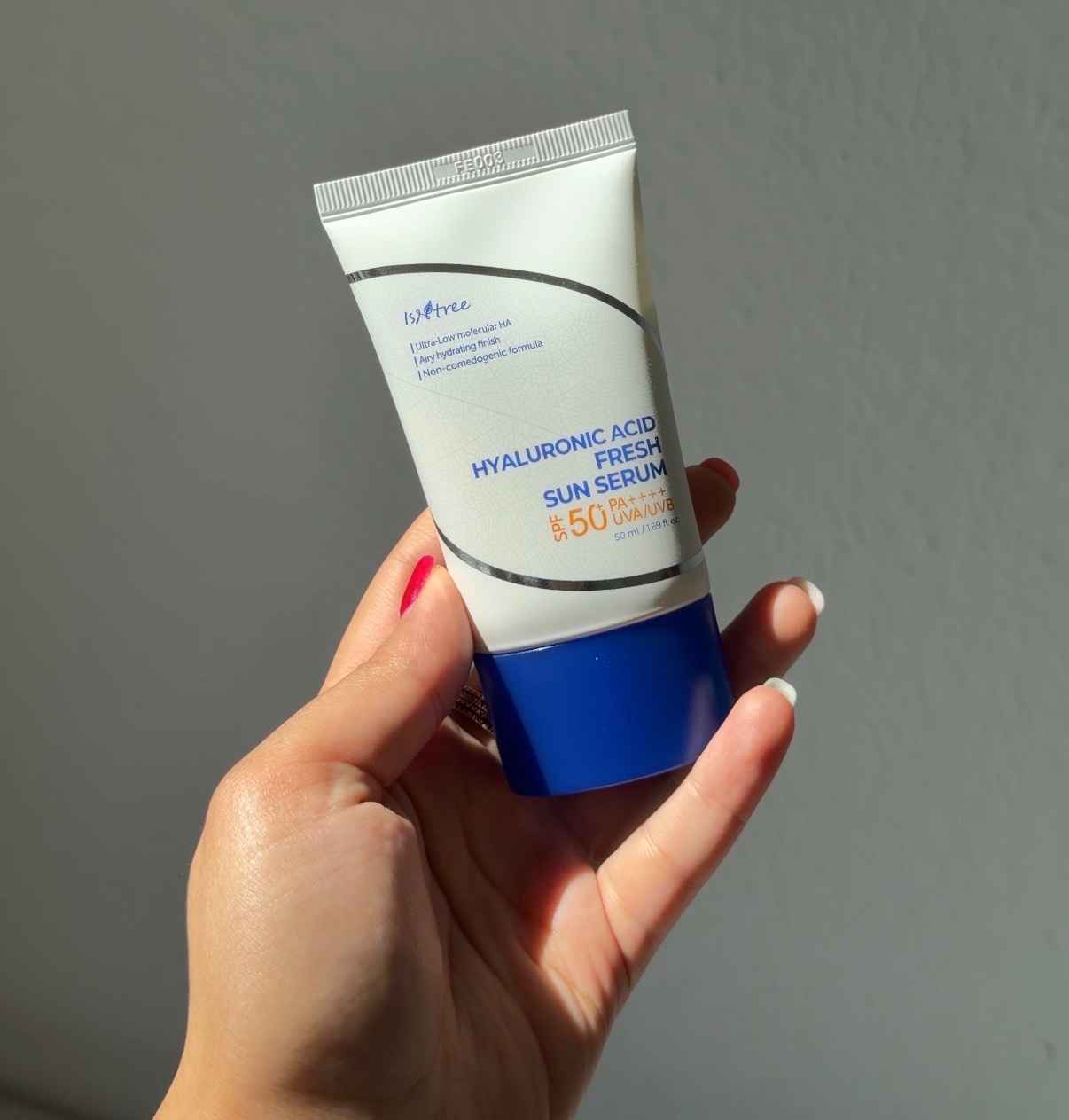 A hand holding a tube of hyaluronic acid fresh sun serum with SPF 50 against a neutral background.