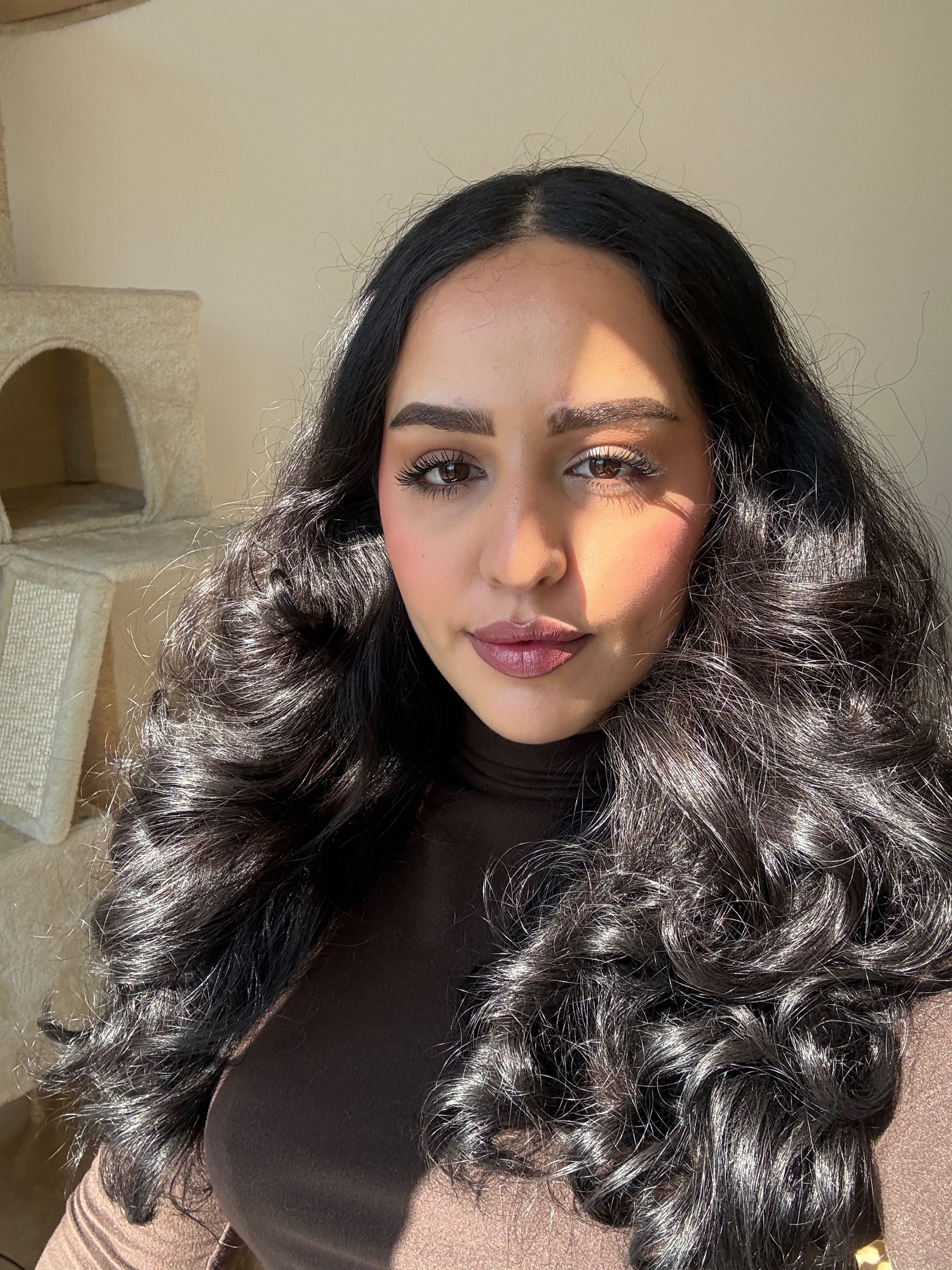 A woman with long, curly black hair poses in soft natural light, showcasing a warm expression and subtle makeup.