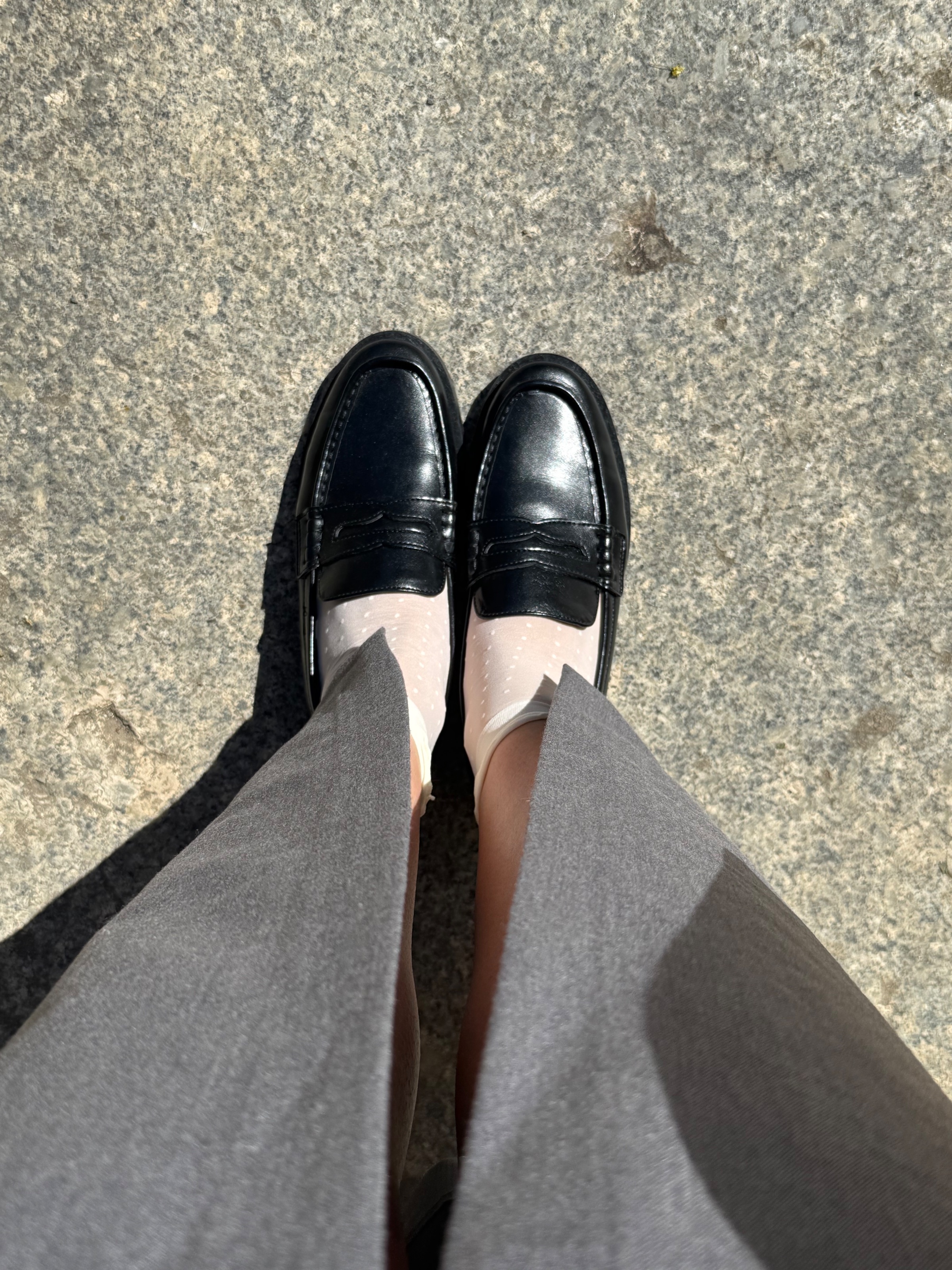 A close-up view of polished black loafers worn with sheer socks, standing on a textured gray surface.