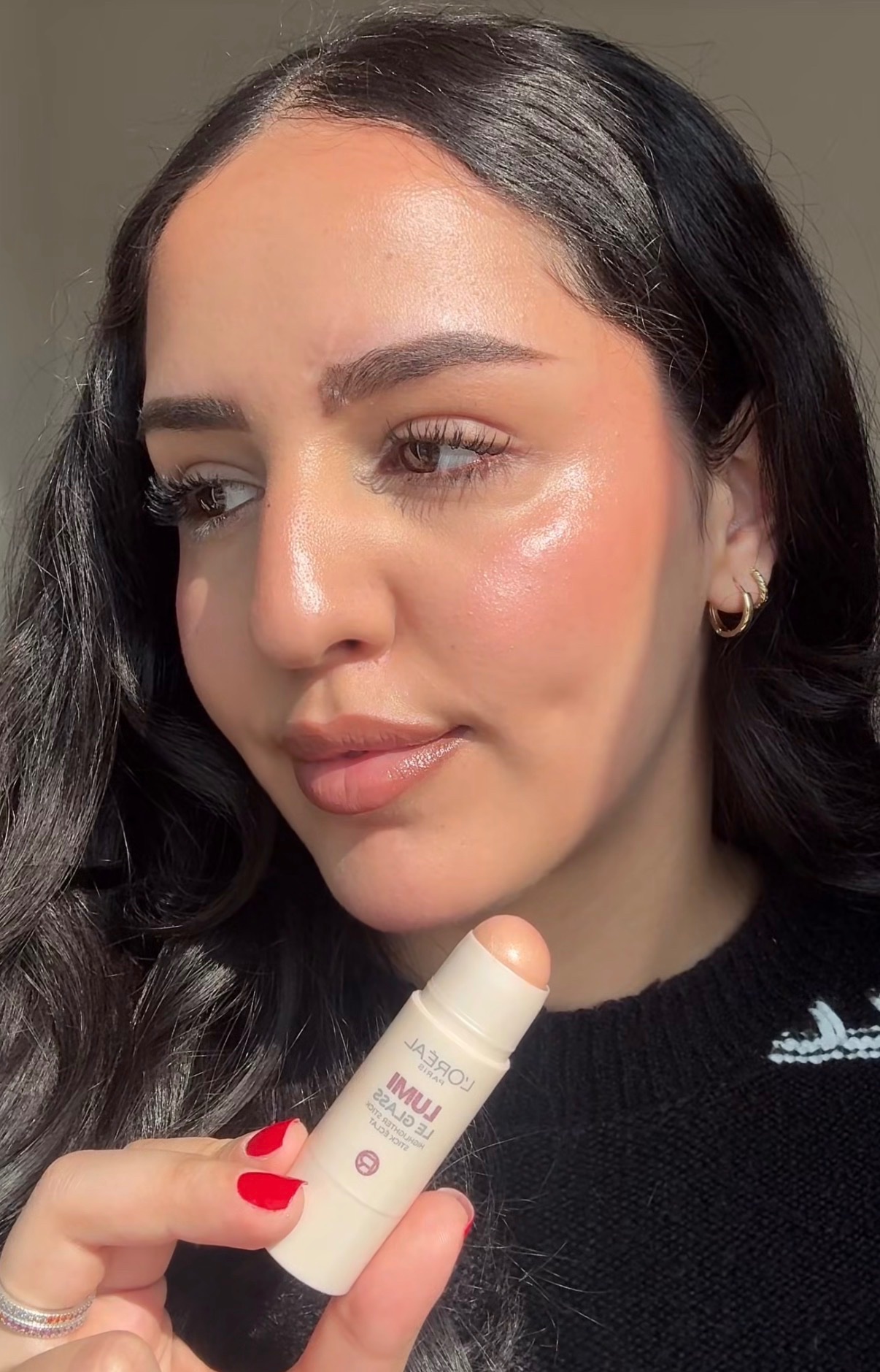 A woman with long, wavy hair holds a makeup stick close to her face, showcasing a natural glow on her skin.