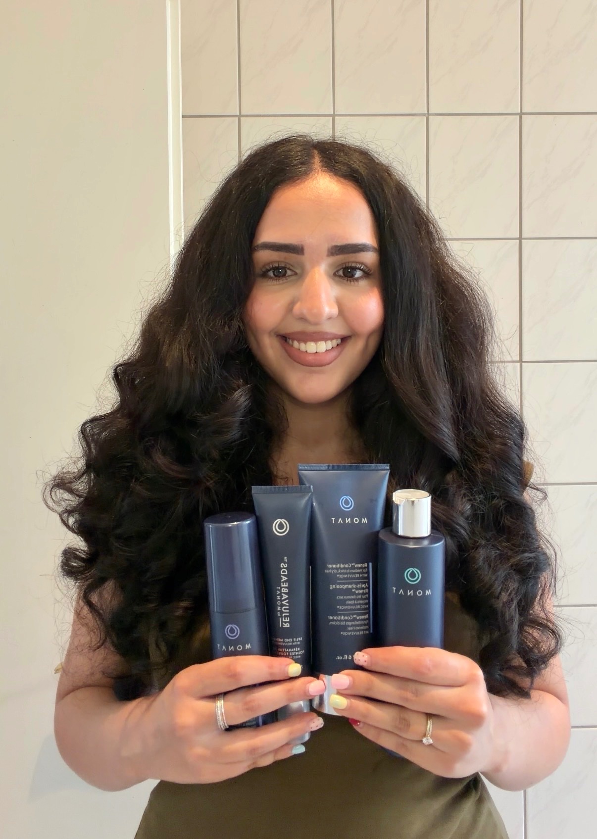 A woman with long, curly hair smiles while holding four skincare products in her hands, showcasing a beauty brand.