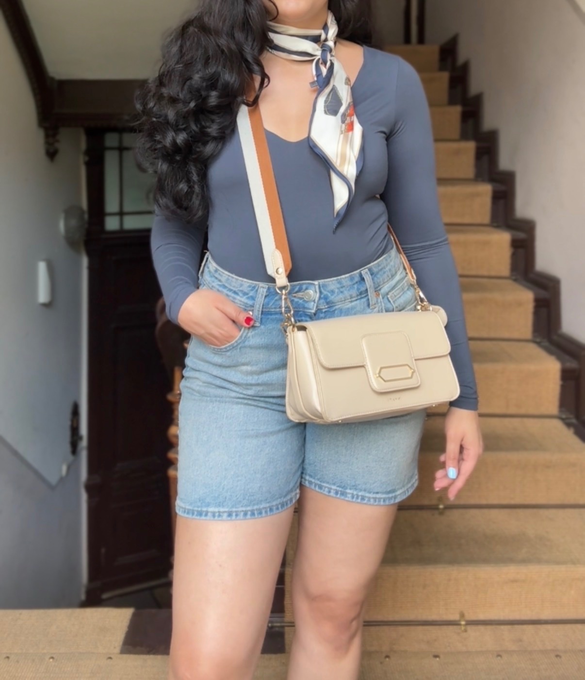 A person stands on a staircase wearing a fitted gray top and high-waisted denim shorts, accessorized with a scarf and a beige handbag.